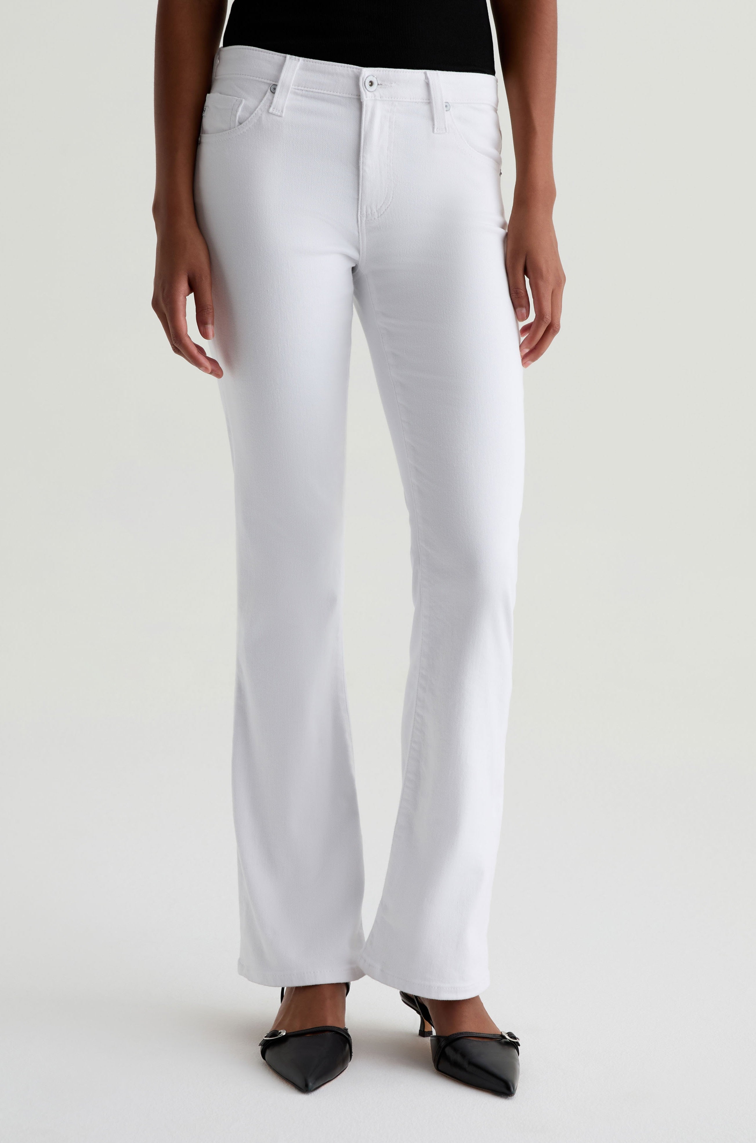 A person wears the AG Jeans Angel white low-rise bootcut jeans with black pointed-toe shoes, standing against a plain light background. Their relaxed arms are at their sides and only part of their upper body is visible.