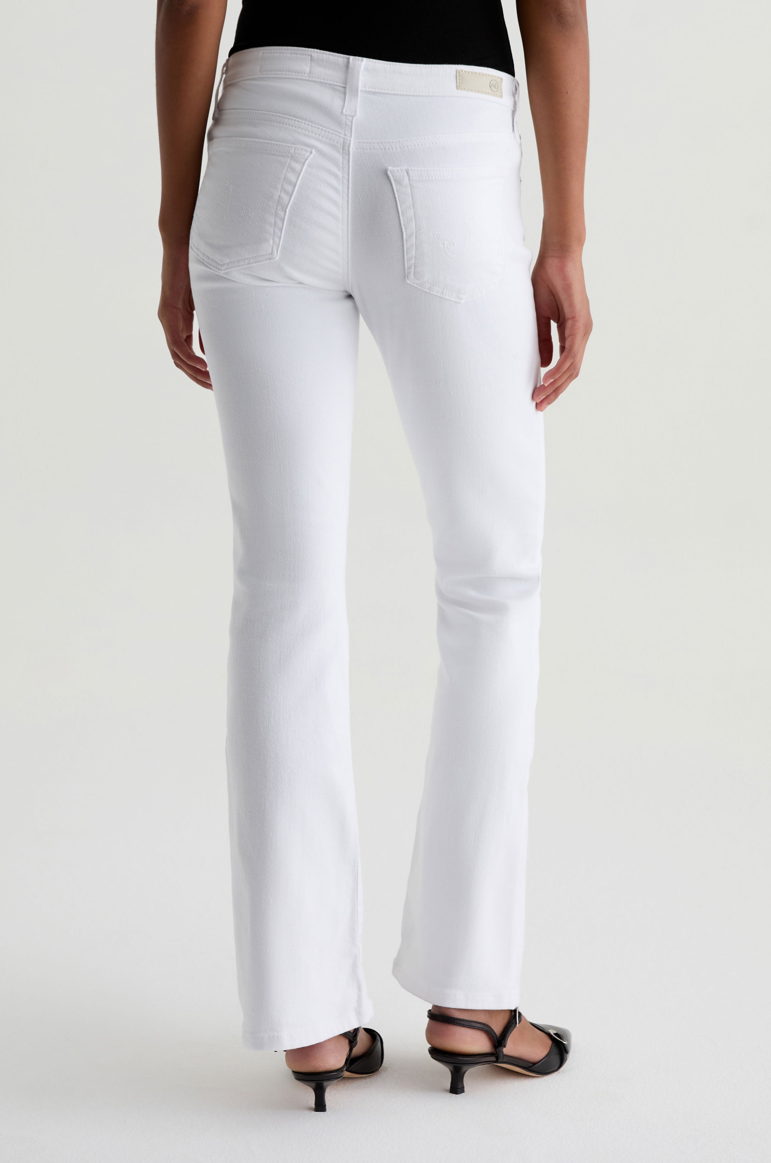A person stands with their back to the camera wearing AG Jeans' Angel white jeans, a black top, and black open-toe heels on a light background.