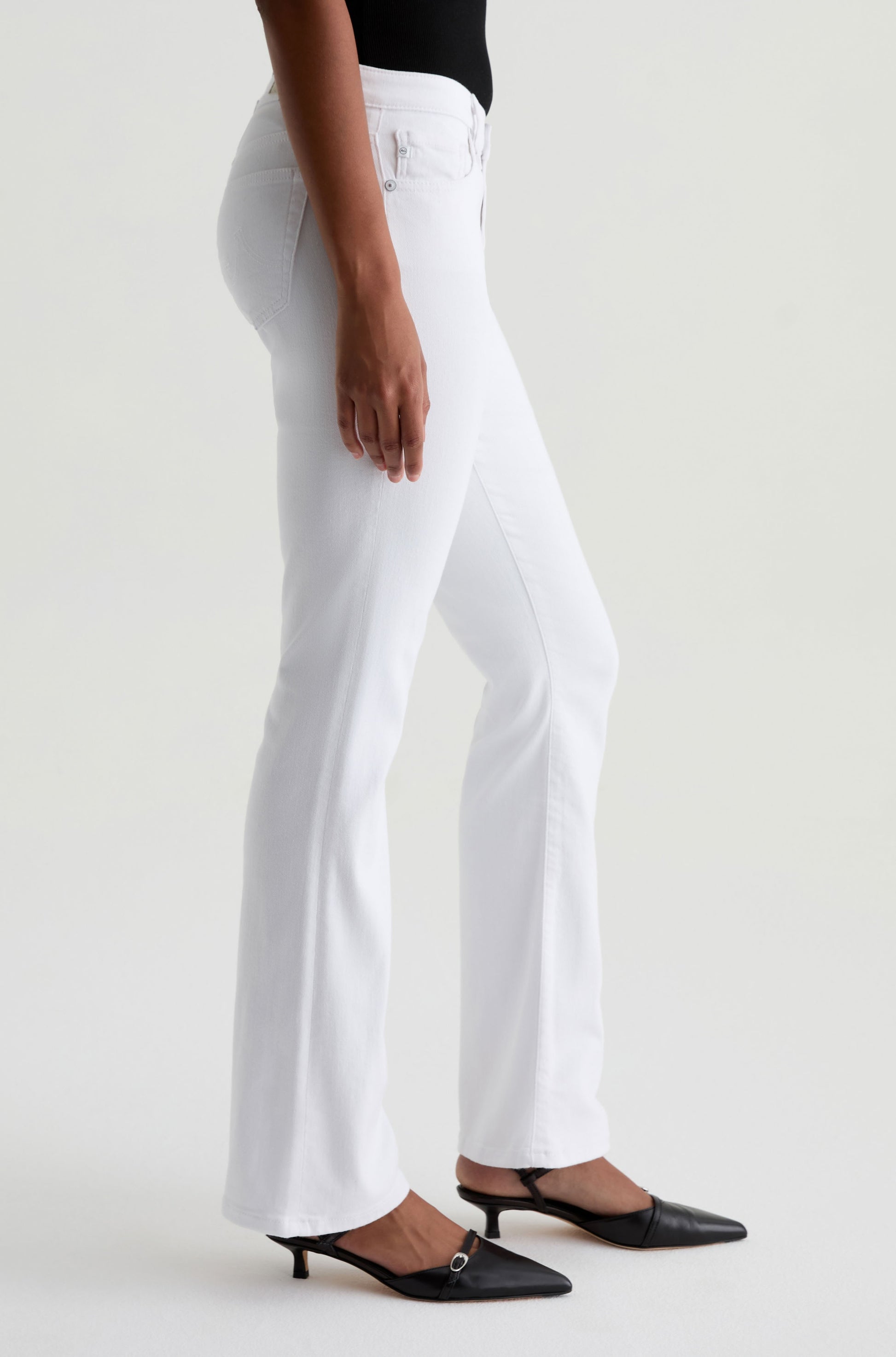 A person wears the AG Jeans Angel white low-rise bootcut jeans, a black sleeveless top, and black low-heeled slingback shoes against a plain light background.