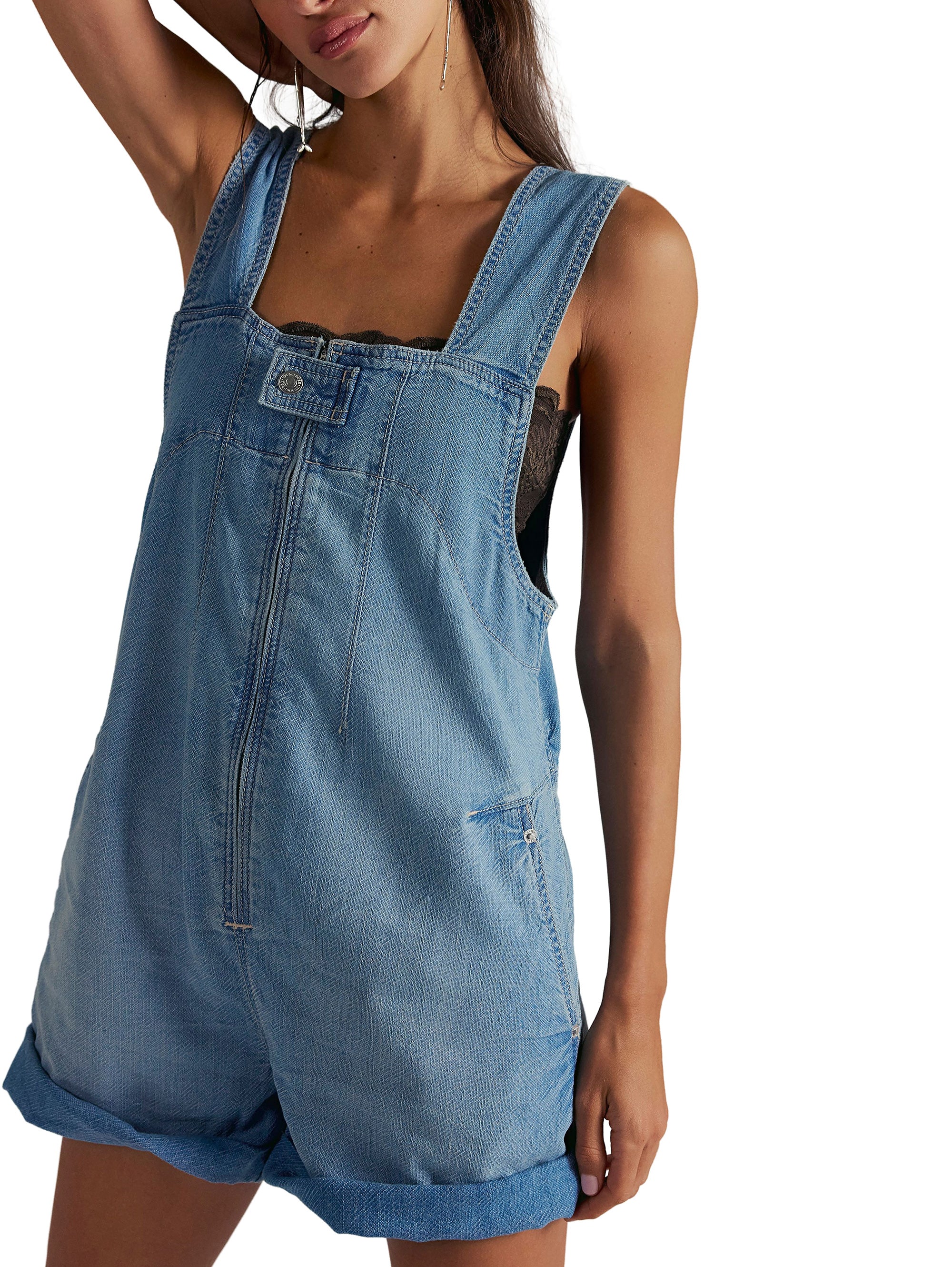 A woman wears the Free People Jude Shortall in medium denim—100% cotton, with wide straps, front pockets, and folded hems. She stands against a white background with her left arm raised; her face is partially out of frame.