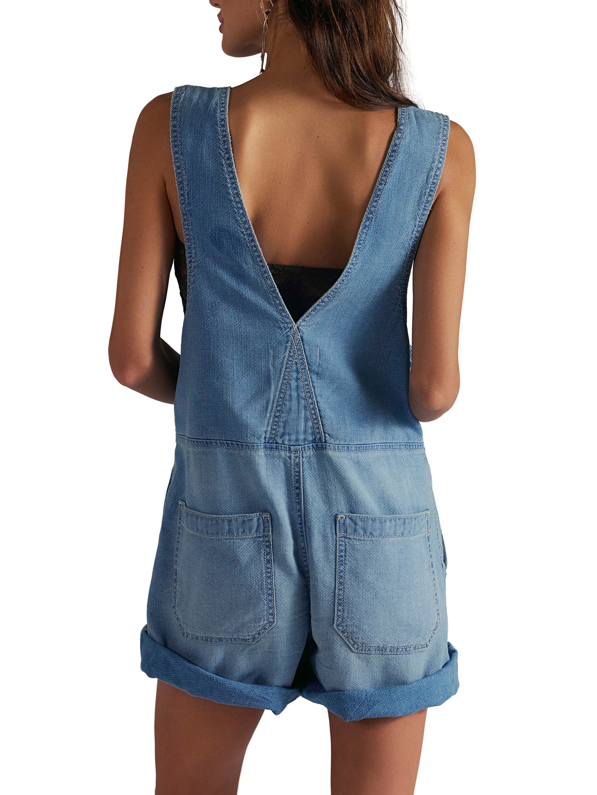 A woman with long brown hair is seen from the back wearing the Free People Jude Shortall, a sleeveless, light blue 100% cotton denim romper featuring a deep V-back, two back pockets, and rolled-up hem shorts.