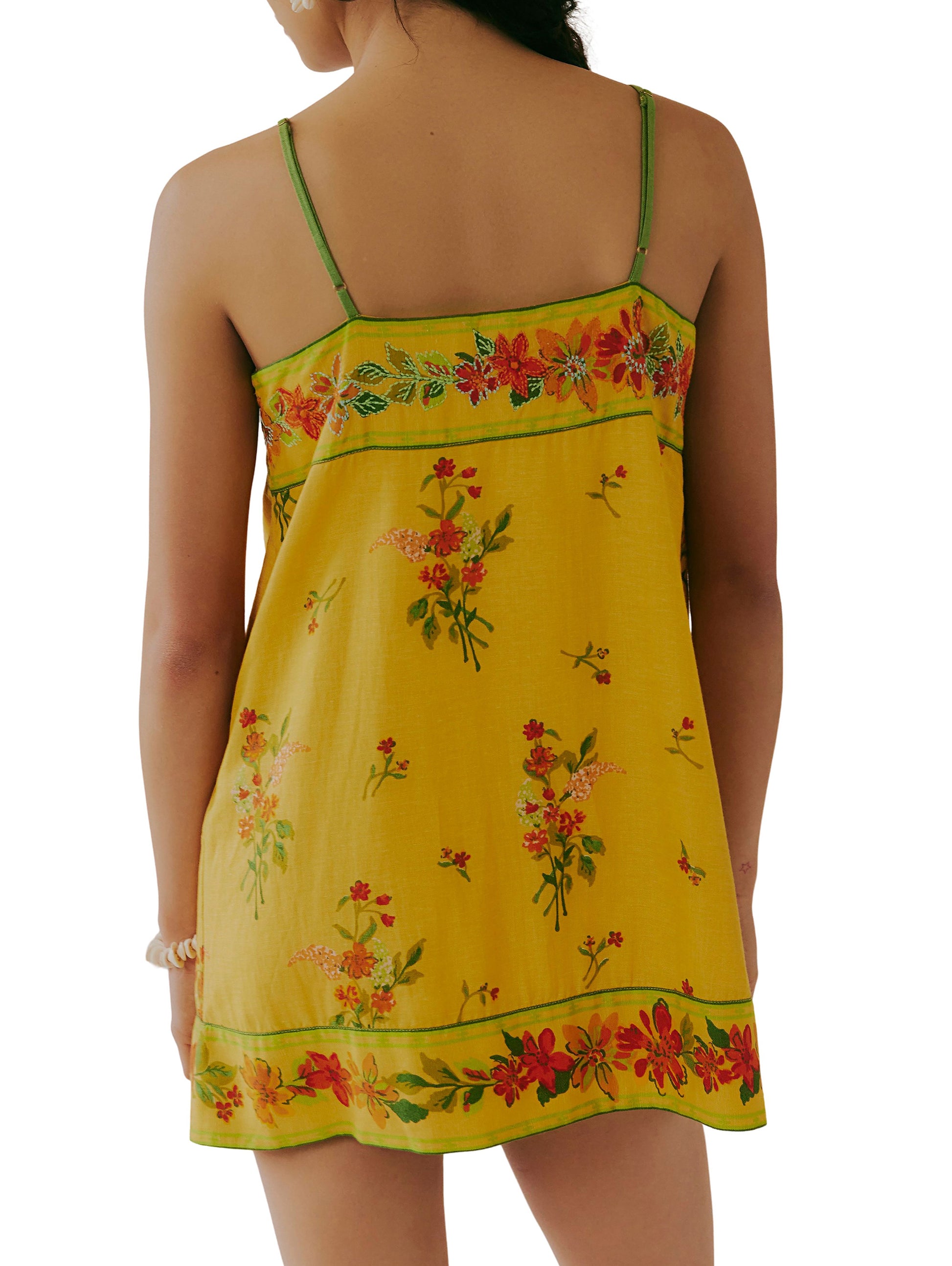 A woman is shown from the back wearing the Free People Bali Take Me With You Mini, a yellow floral print dress with adjustable shoulder straps and colorful flower designs throughout.