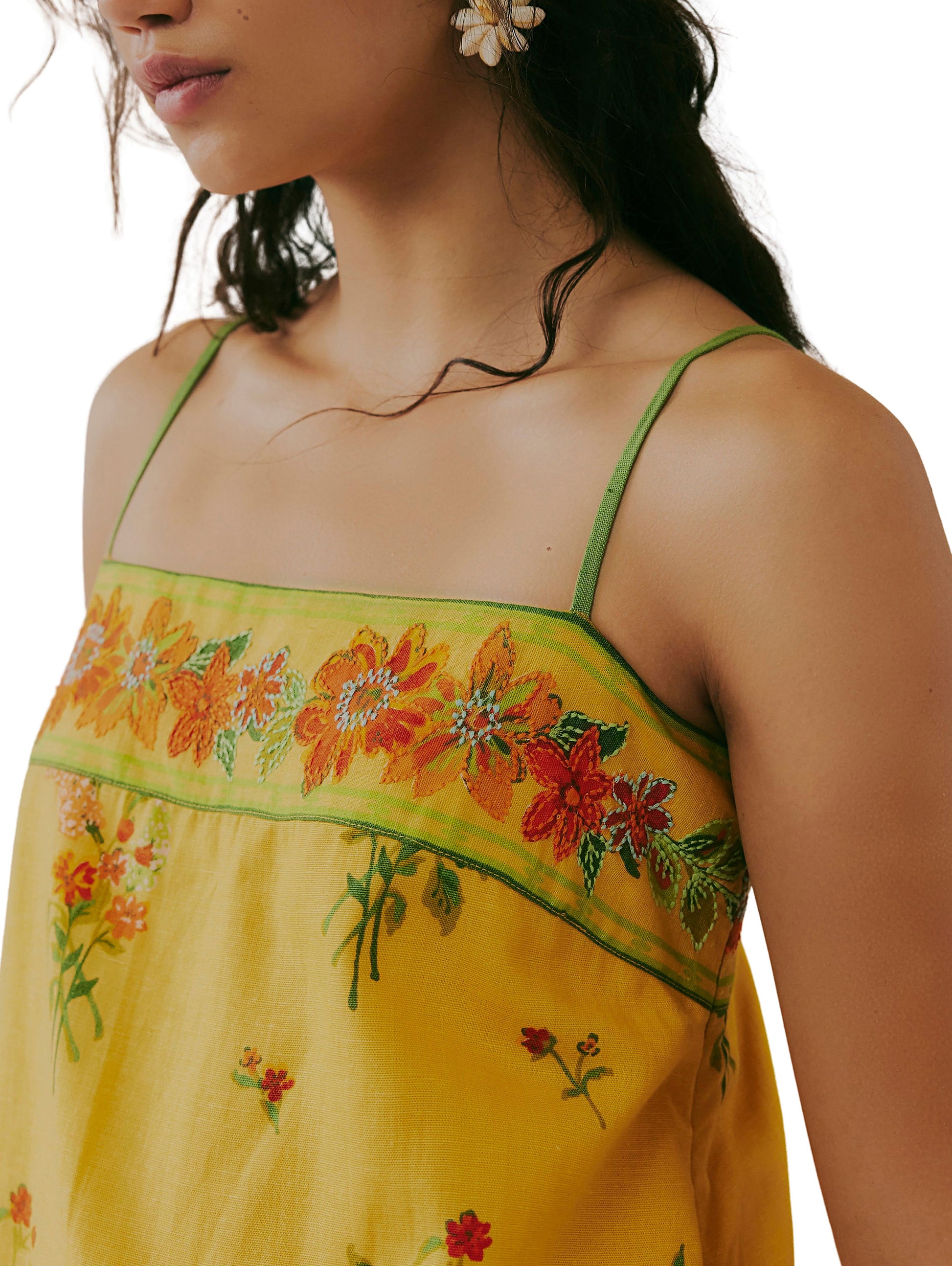 A woman in the Free People Bali Take Me With You Mini, a yellow floral print dress with adjustable straps and red, orange, and green embroidered details; she wears floral earrings and has long dark hair.