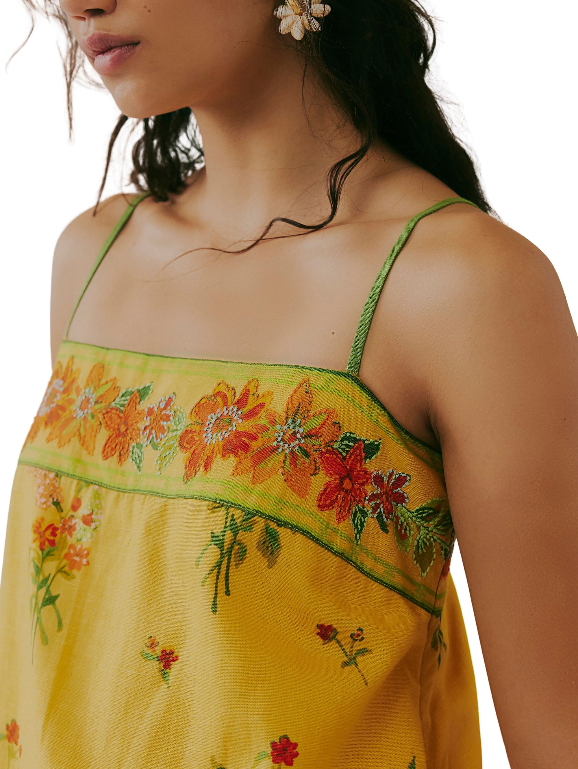 A woman in the Free People Bali Take Me With You Mini, a yellow floral print dress with adjustable straps and red, orange, and green embroidered details; she wears floral earrings and has long dark hair.