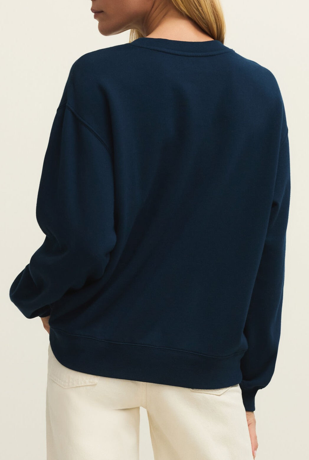 A person is shown from behind wearing the Z Supply Yachty Boyfriend Sweatshirt in dark blue, made from midweight cotton with a crewneck and long sleeves, paired with off-white pants against a plain light background.
