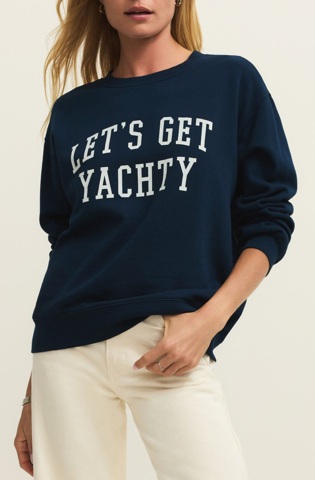 A woman models the Z Supply Yachty Boyfriend Sweatshirt—a navy blue midweight cotton crewneck with white “LETS GET YACHTY” text—paired with white pants, against a plain background.
