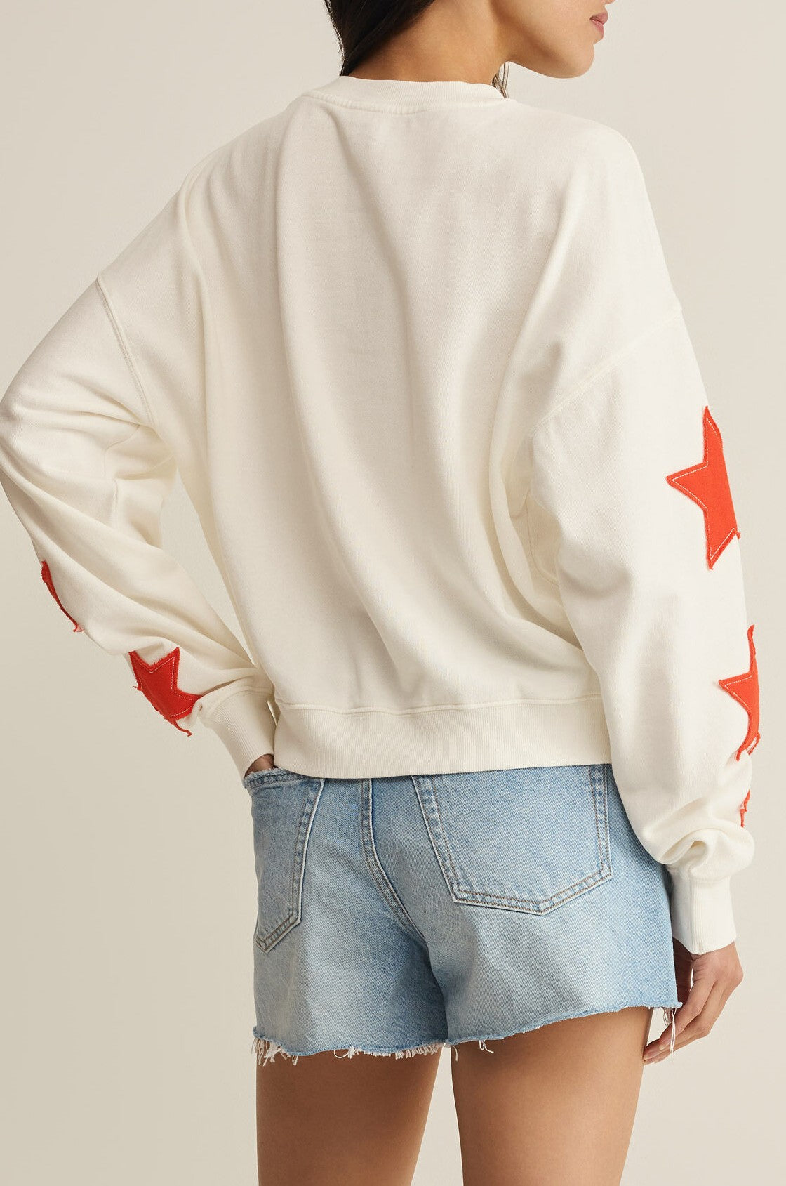 A woman wears the Z Supply Stars First Date Sweatshirt—100% cotton, white with red stars on the sleeves—and light blue denim shorts, shown from the back.