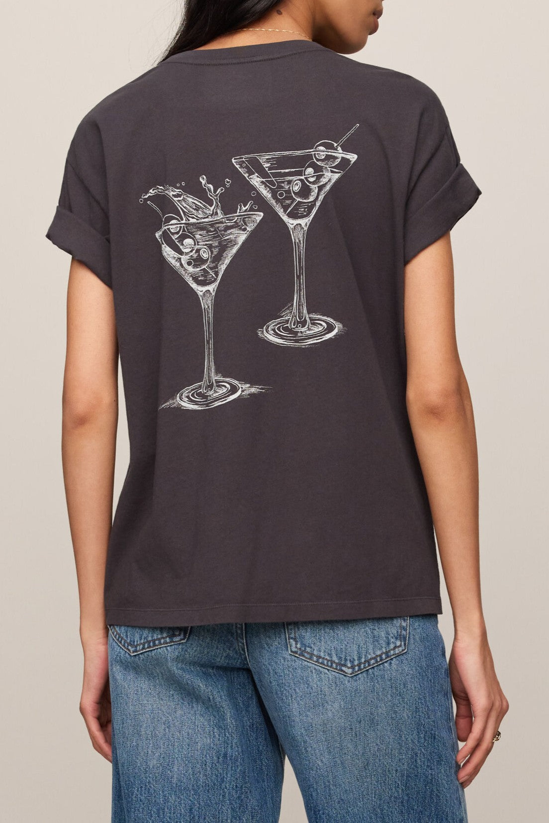 A person wearing the Z Supply Martini Boyfriend Tee— a regular fit, 100% cotton dark T-shirt featuring white line drawings of two martini glasses on the back—paired with blue jeans.