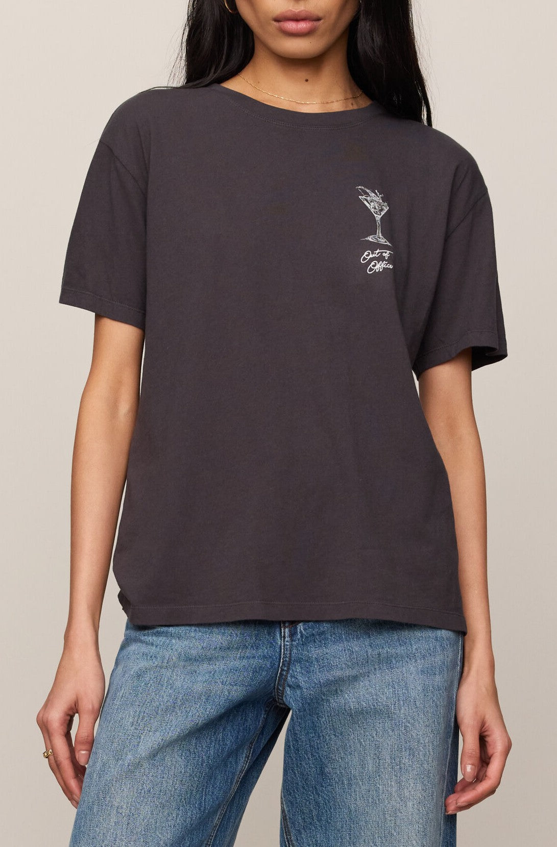 A person wearing the Z Supply Martini Boyfriend Tee in BLACK SAND—a loose-fitting, regular fit crew neck t-shirt made from 100% cotton with a small white cocktail glass graphic and text on the left chest—paired with blue jeans.