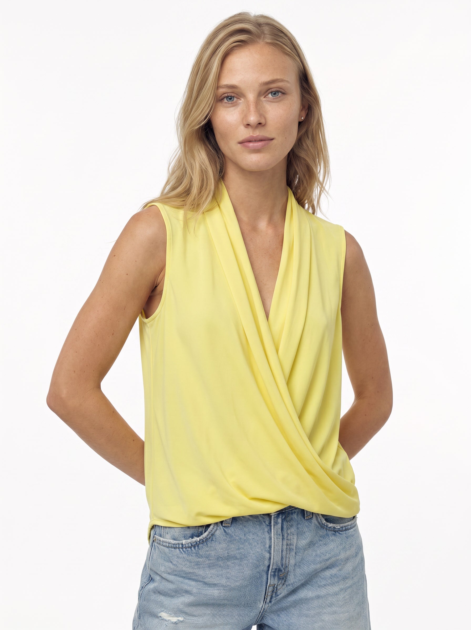 A blonde woman wears the Georgie Gwen Sleeveless Wrap Top in yellow with light blue denim shorts, standing against a plain white background with her hands behind her back.
