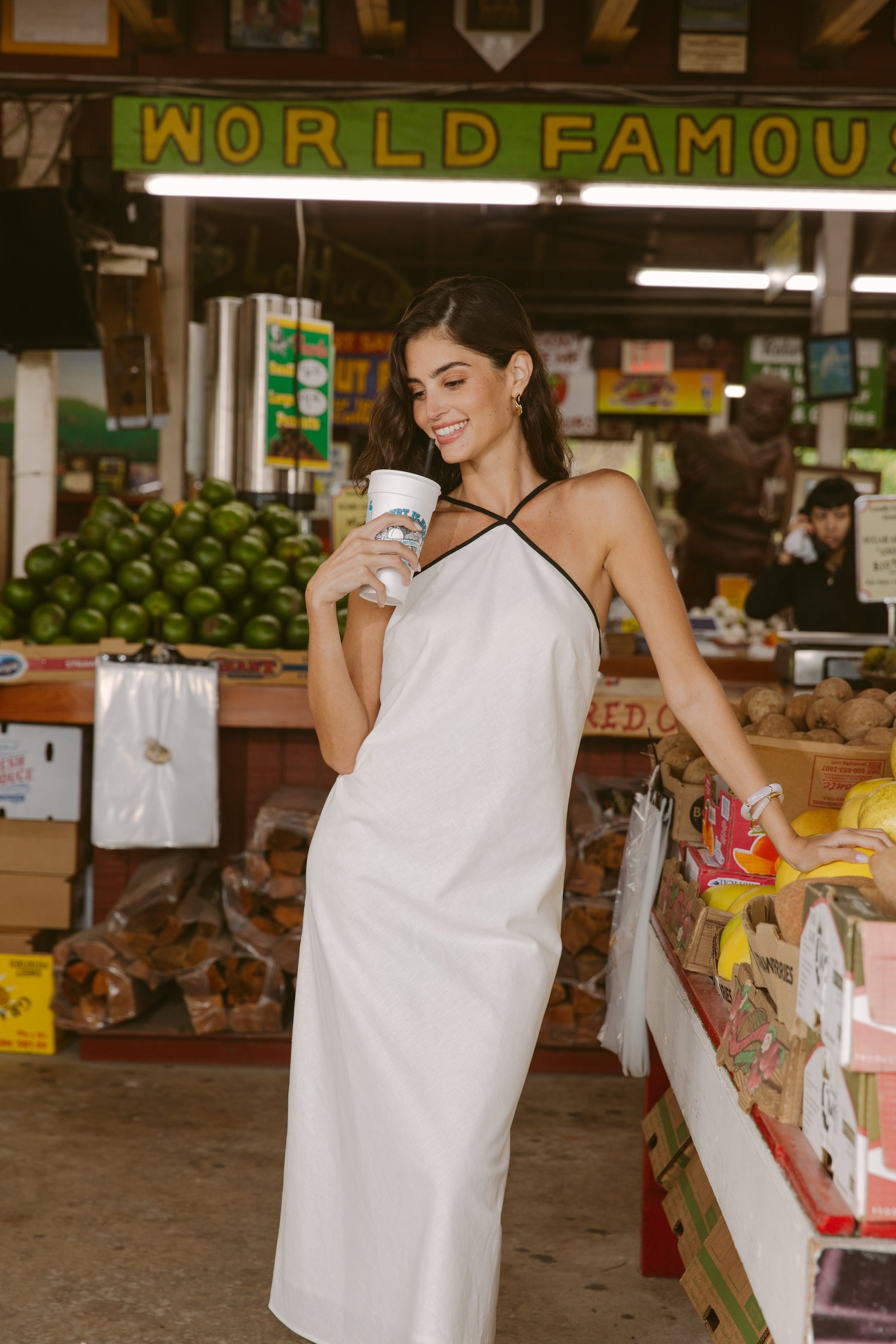 A woman wearing the Georgie Margot Linen Midi Dress smiles while holding a drink in a lively market, with stacks of fruit and a "WORLD FAMOUS" sign behind her.