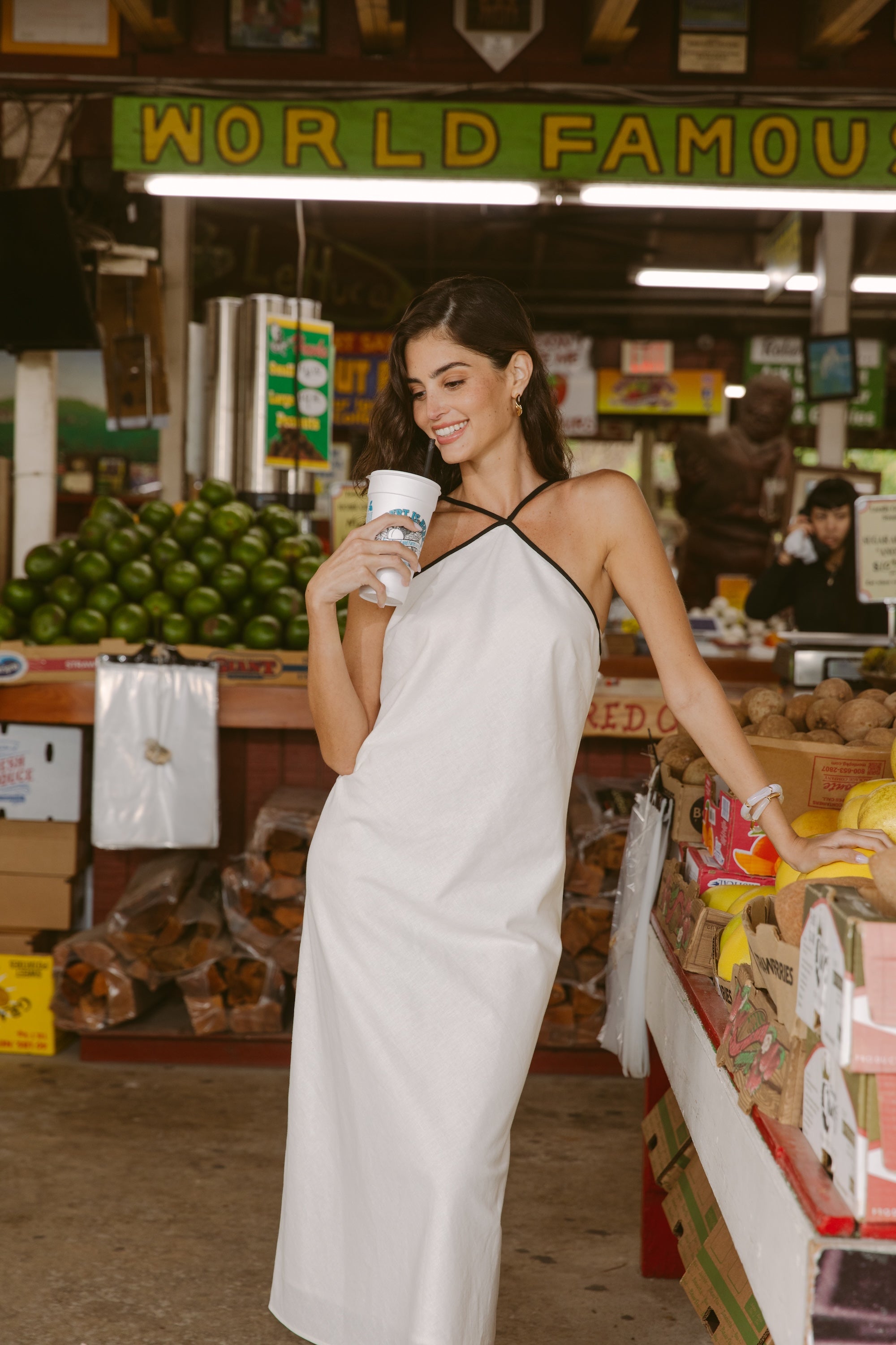 A woman wearing the Georgie Margot Linen Midi Dress smiles while holding a drink in a lively market, with stacks of fruit and a "WORLD FAMOUS" sign behind her.