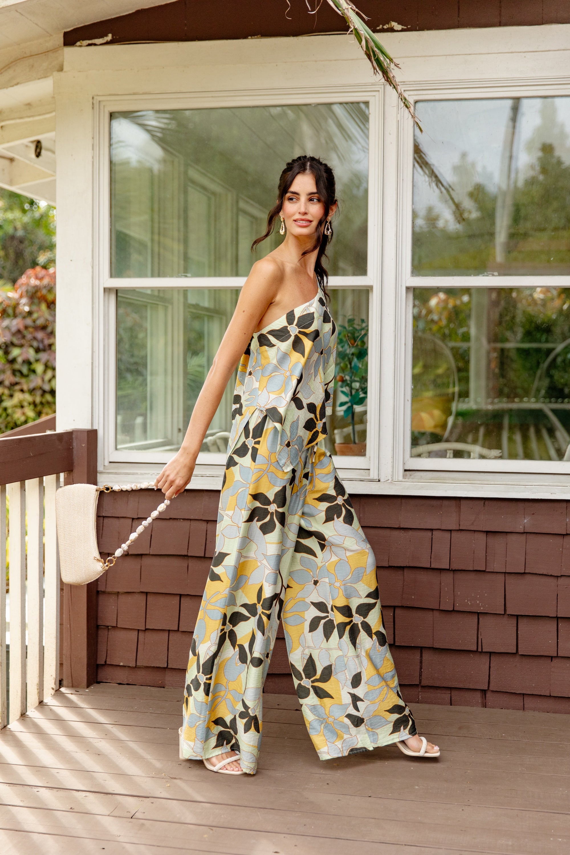 A woman wearing The Nora Set Top by Georgie stands on a porch, holding a woven bag and smiling slightly. She looks to the side, with trees and windows in the background.
