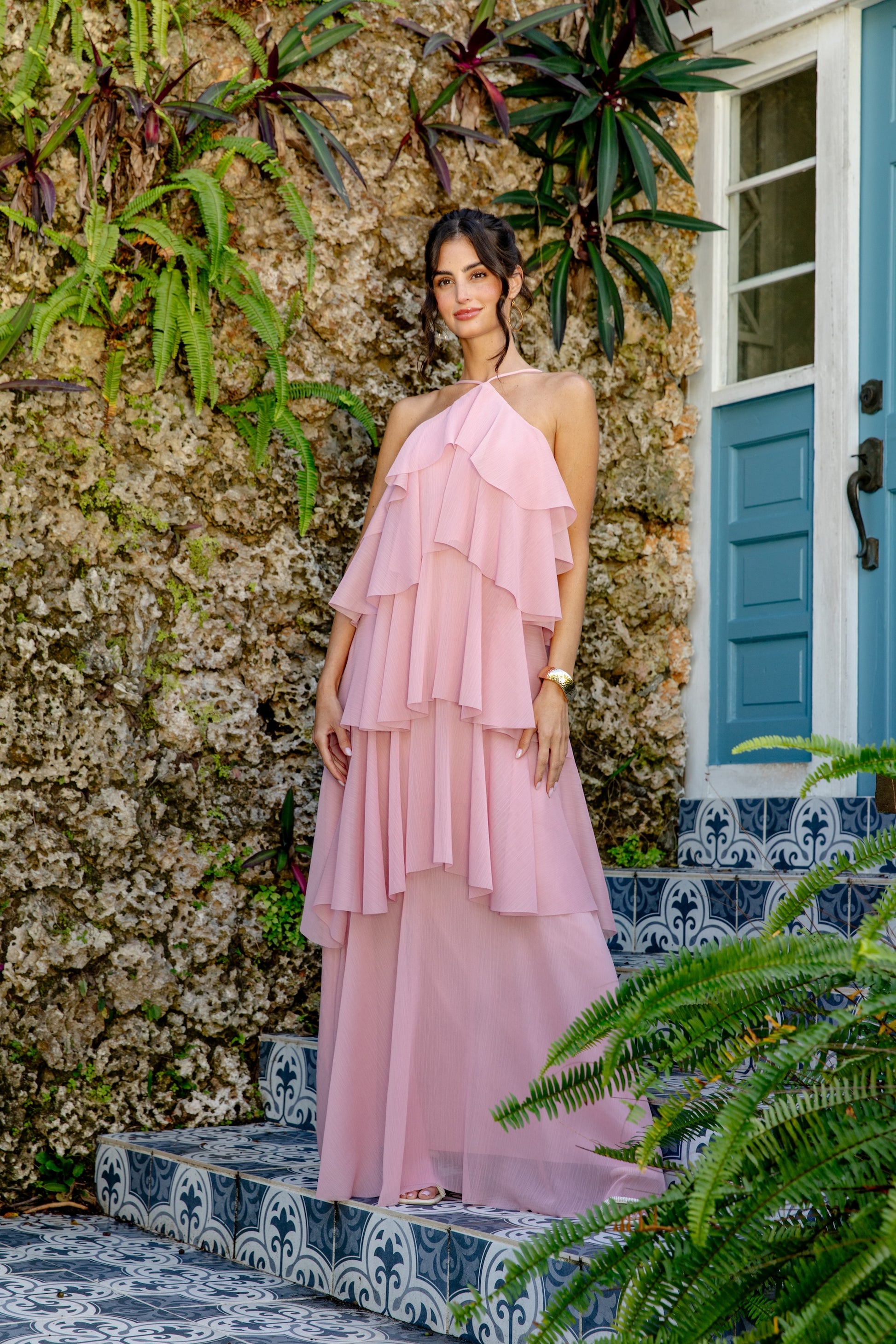 A woman in the Georgie Ophelia Ruffle Tiered Maxi Dress stands on patterned blue-tiled steps beside a stone wall with lush green plants, near a blue door with glass panels.