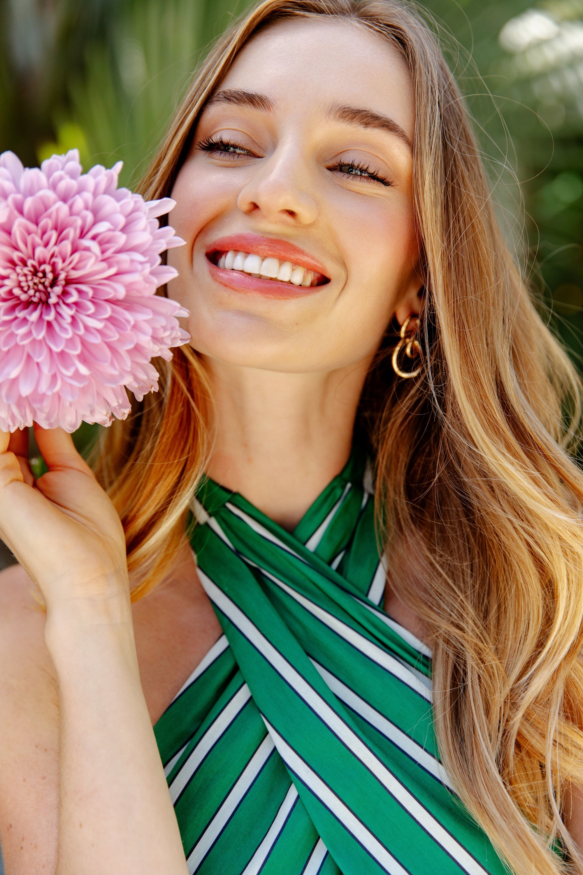 A smiling woman with long blonde hair holds a pink flower near her face, wearing the Georgie Parker Halter Maxi Dress with green and white stripes and gold hoop earrings, outdoors with blurred greenery in the background.