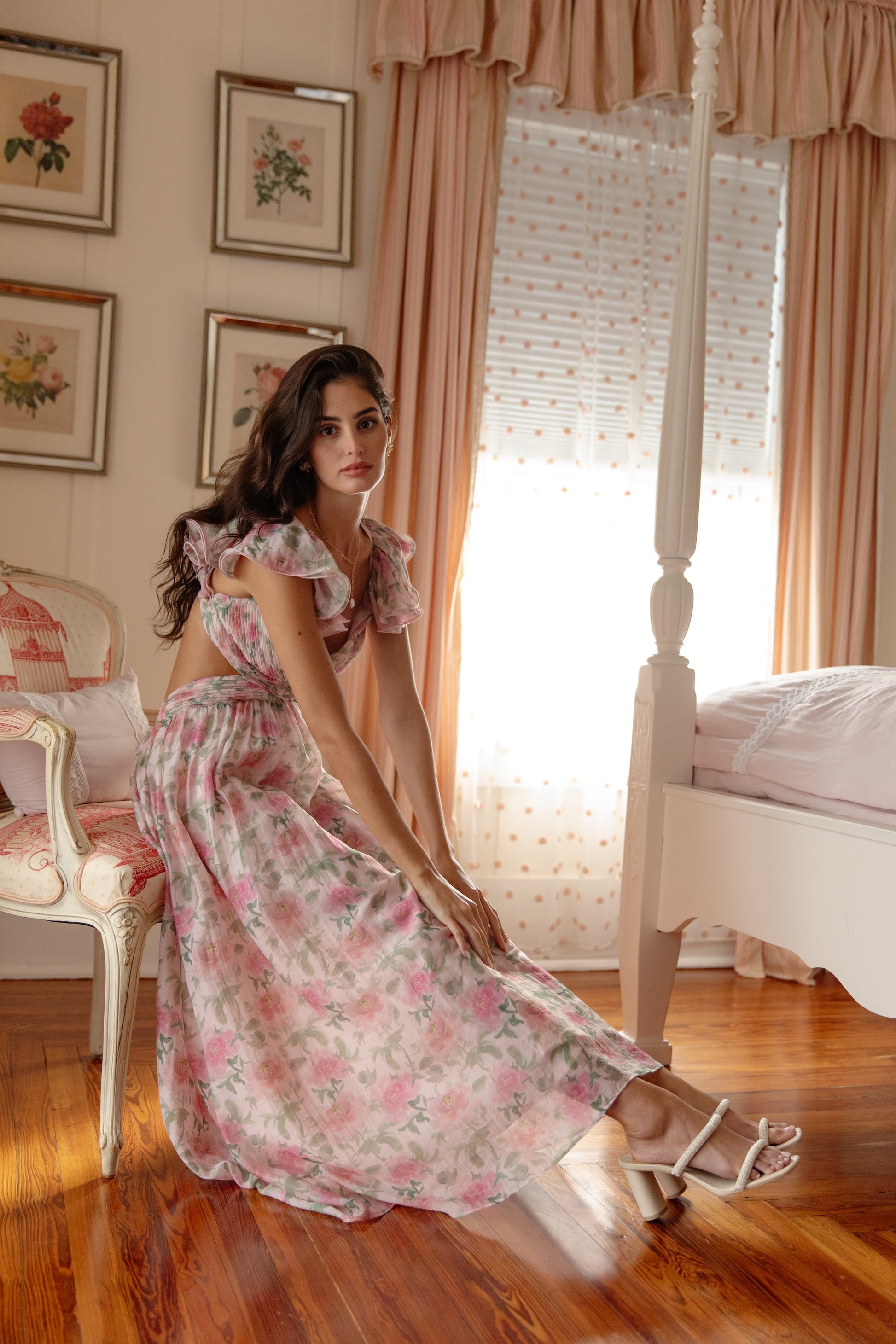 A woman in the Georgie Rosalie Flutter Sleeve Maxi Dress sits by a four-poster bed in a sunlit, vintage-inspired room with floral art and pink curtains—an elegant look for any special occasion.