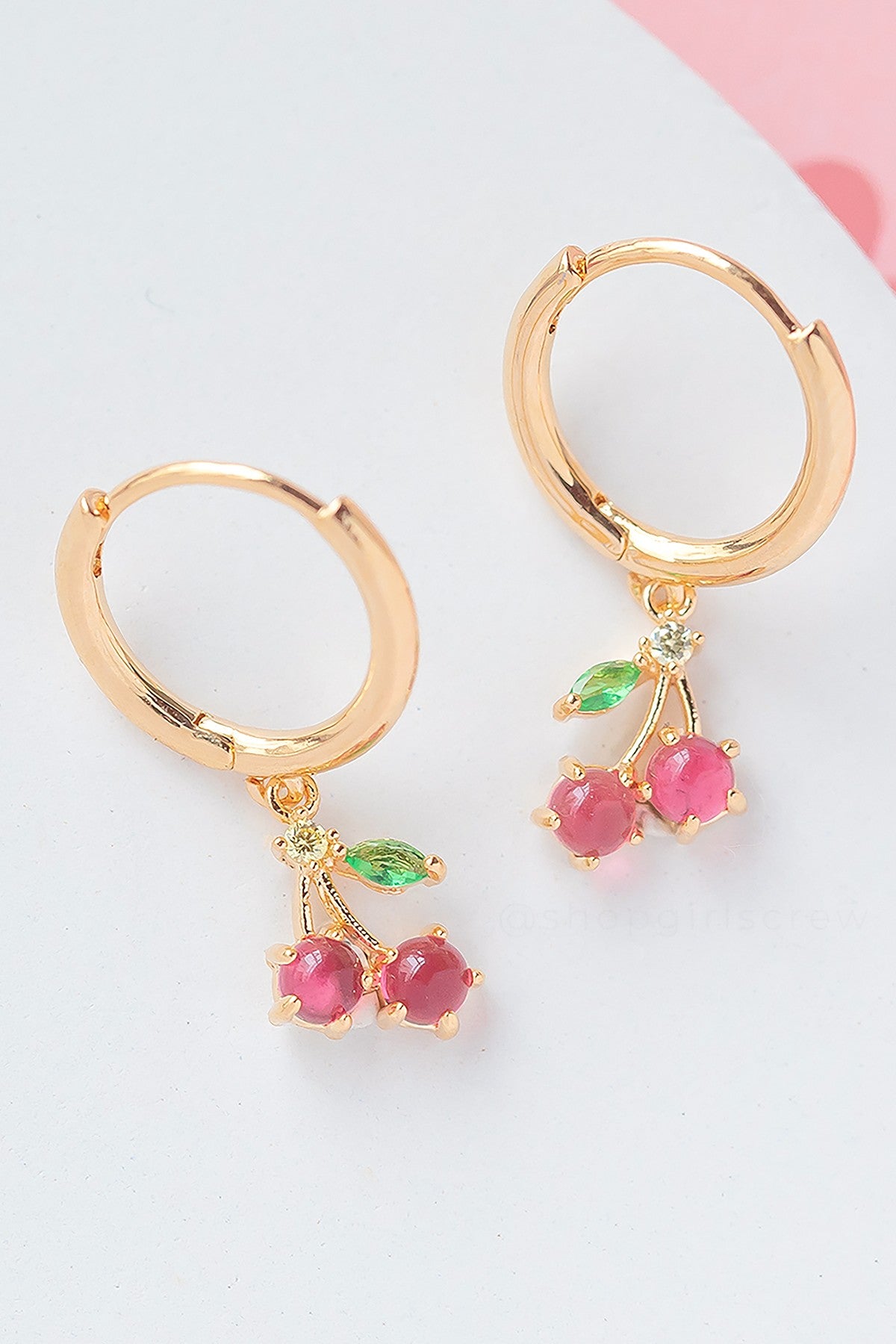 The Cherry Sweet Mini Hoops by Girls Crew are pink 18k gold plated earrings with cherry-shaped charms and green leaf accents, shown on a white surface.