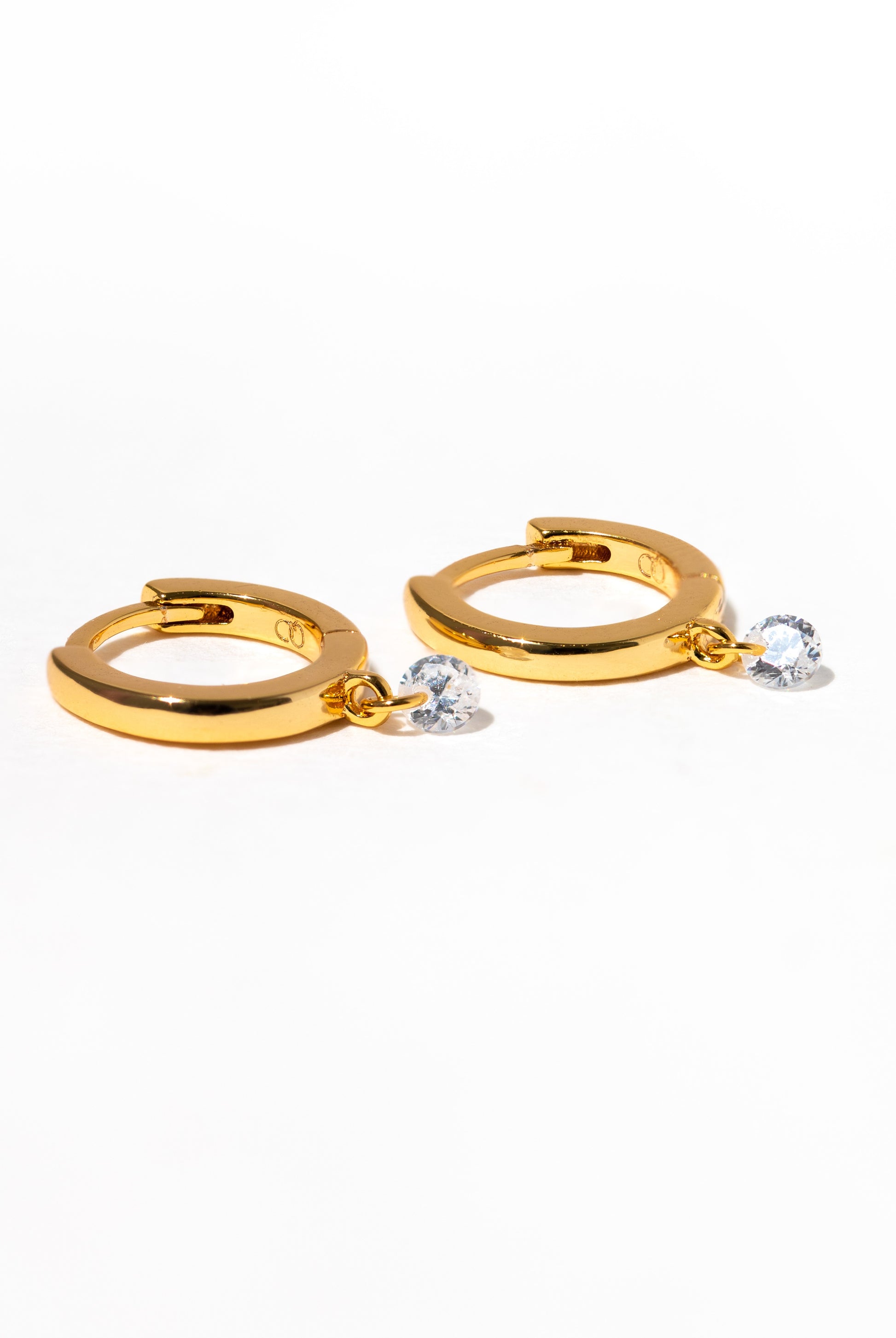 The Dew You Hoops by Girls Crew are 18k gold plated earrings, each with a dangling clear heart-shaped cubic zirconia, shown on a plain white background.