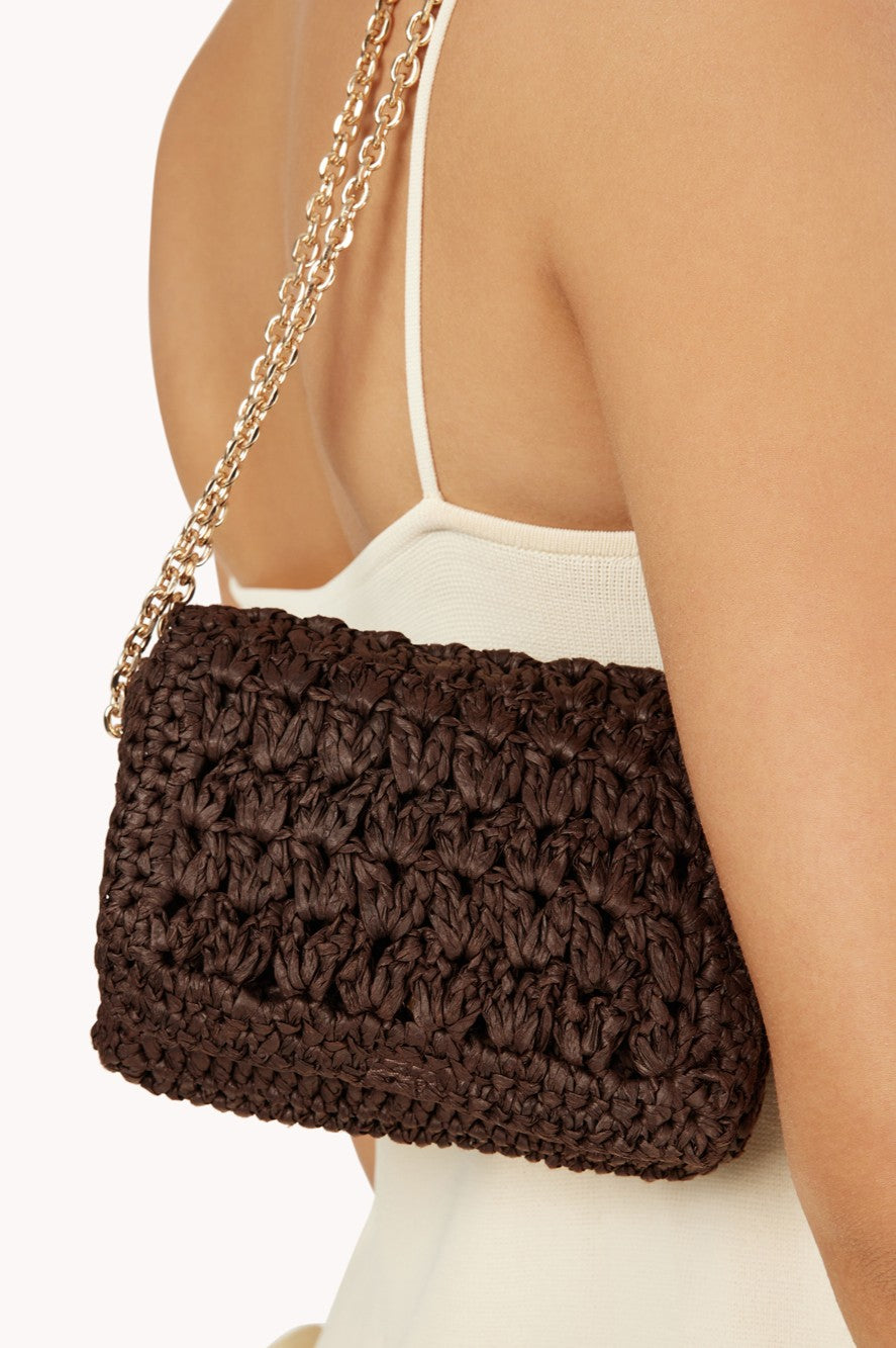 A person wearing a cream-colored top carries the Billini Hayworth Crossbody Bag, a chocolate brown raffia shoulder bag with a gold chain strap.