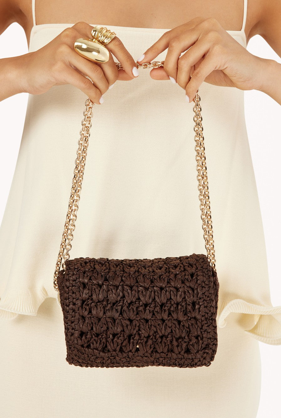 A woman in a cream dress holds the Billini Hayworth Crossbody Bag, featuring chocolate brown raffia and a gold chain strap. Only her torso and hands with a large gold ring are visible.