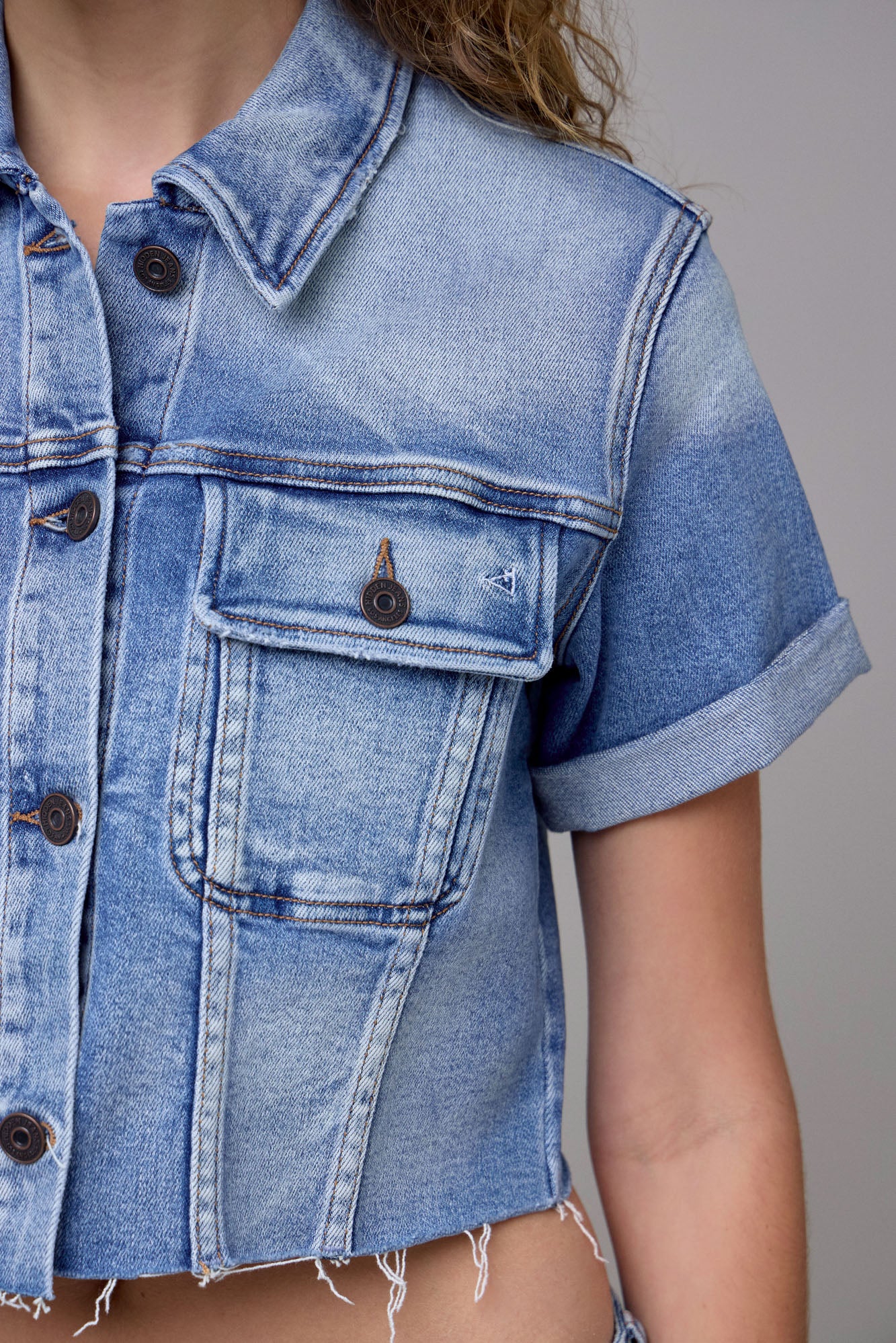 A close-up shows someone wearing the Hidden Jeans Cropped Trucker Jacket—a medium wash, short-sleeve denim piece with frayed edges, a front pocket, and brown buttons. Part of their arm and hair is visible.
