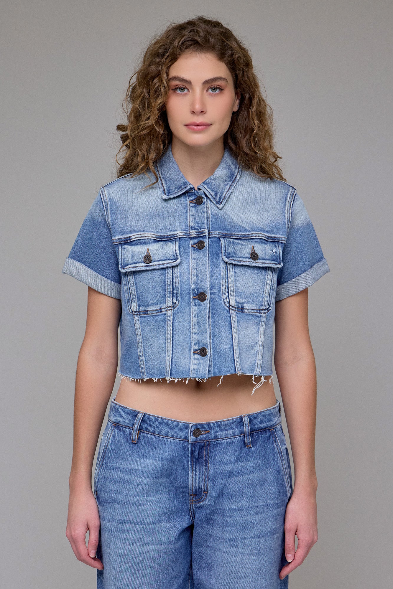 A young woman with curly hair models the Hidden Jeans Cropped Trucker Jacket, featuring a medium wash and rolled cuffs, paired with matching high-waisted jeans against a plain gray background.