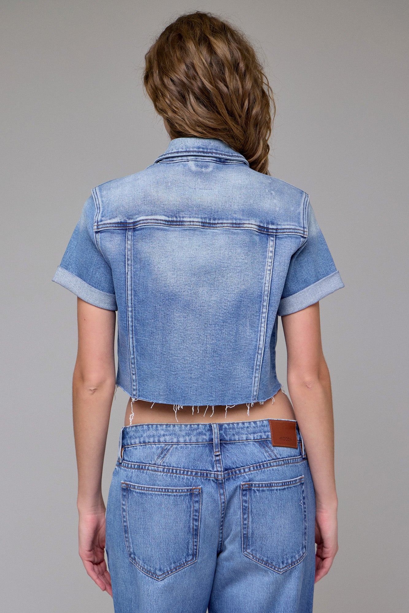 A woman with wavy brown hair is seen from the back against a plain gray background, wearing the Hidden Jeans Cropped Trucker Jacket in light blue with medium wash, paired with high-waisted blue jeans.