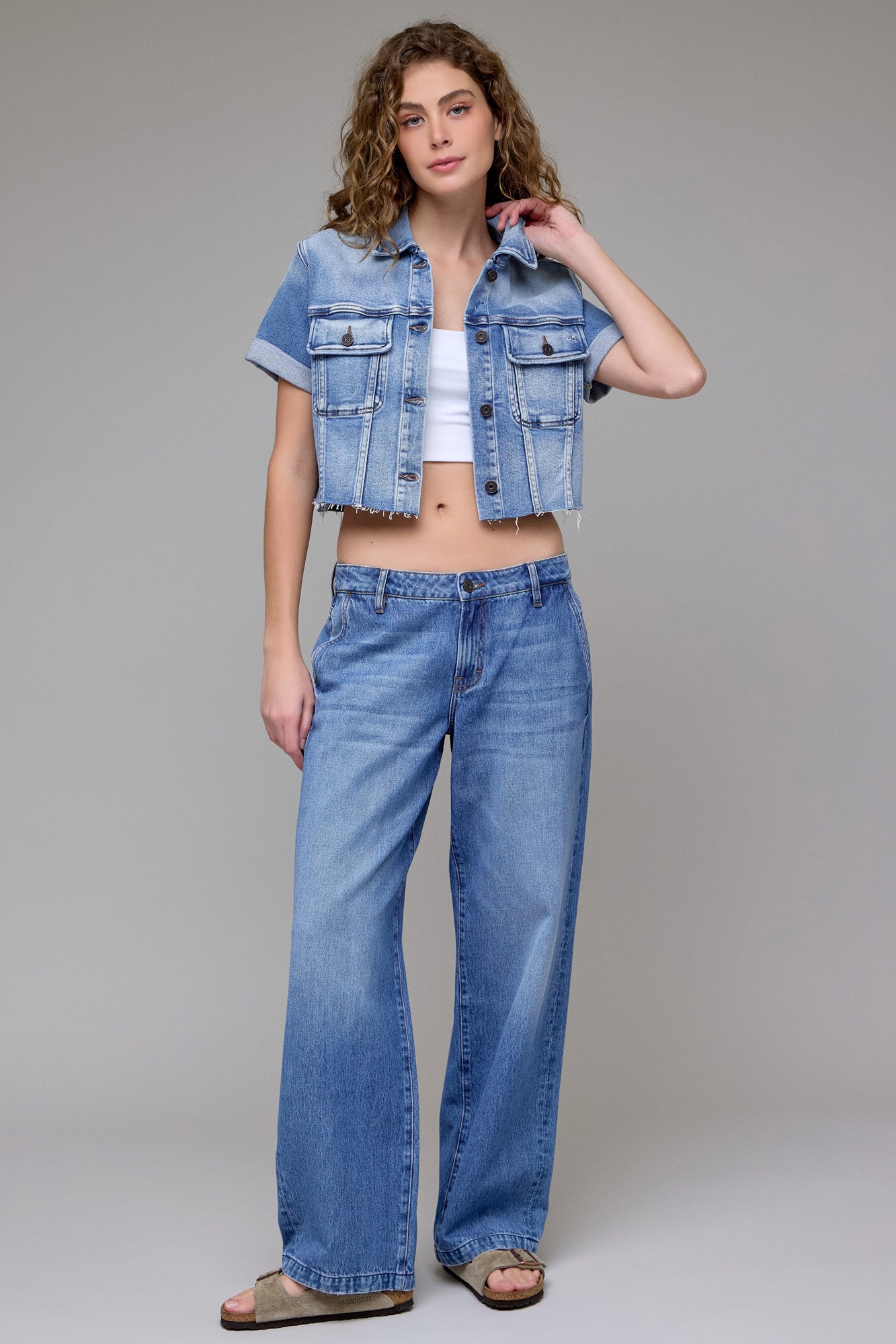 A woman with curly hair wears the Hidden Jeans Cropped Trucker Jacket in a medium wash over a white crop top, paired with loose blue jeans and sandals. She stands facing forward, touching her jacket against a plain gray background.