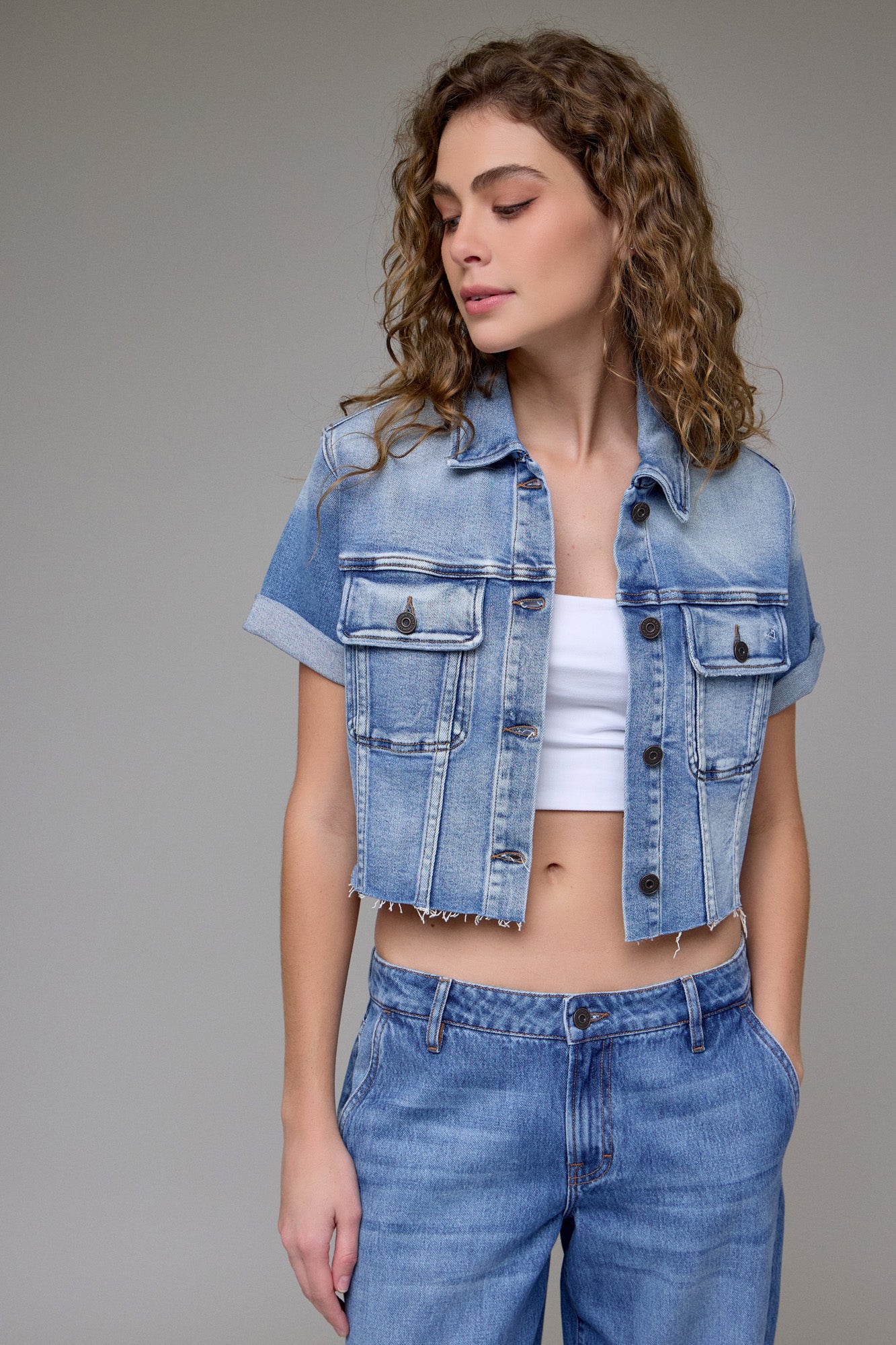 A young woman wears the Hidden Jeans Cropped Trucker Jacket in medium denim over a white crop top and high-waisted jeans, posing with one hand in her pocket and looking slightly to the side.