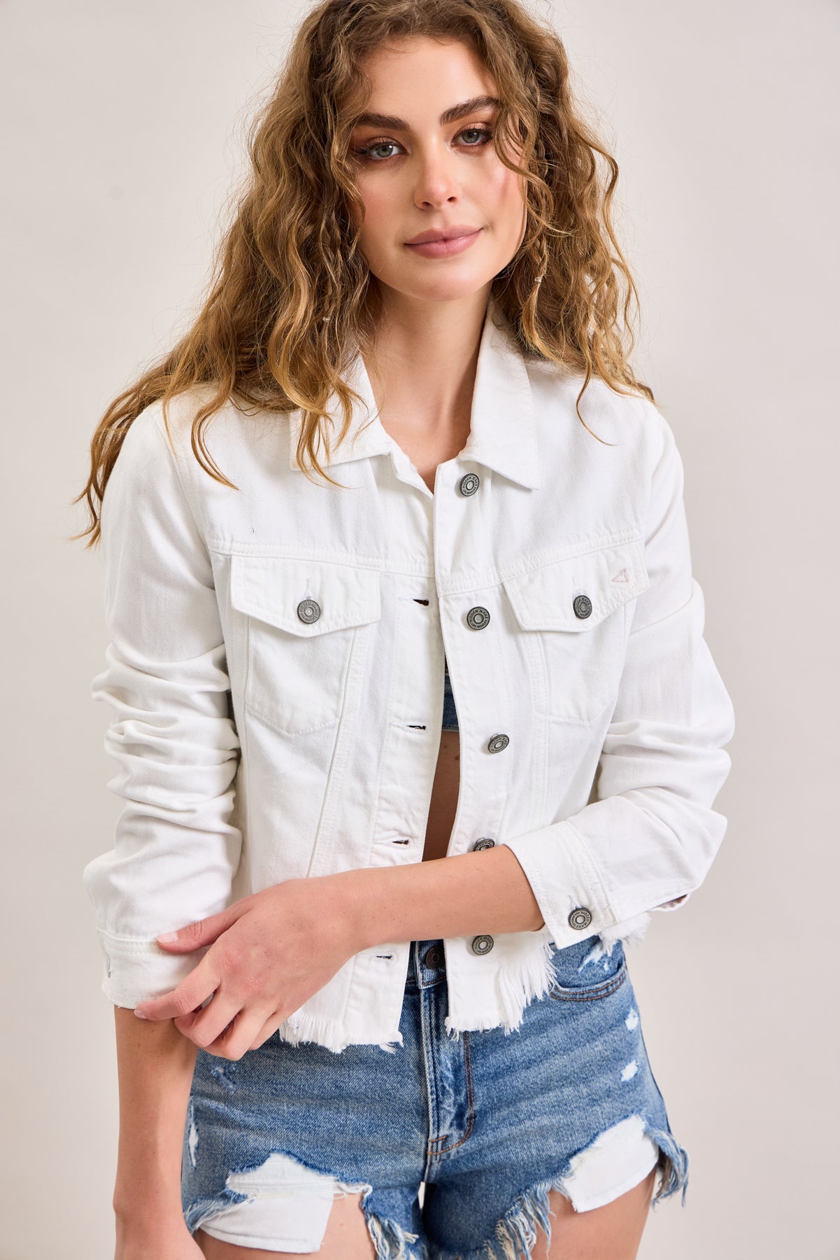 A young woman with curly blonde hair wears the Hidden Jeans Raw Hem Denim Jacket paired with distressed denim shorts.