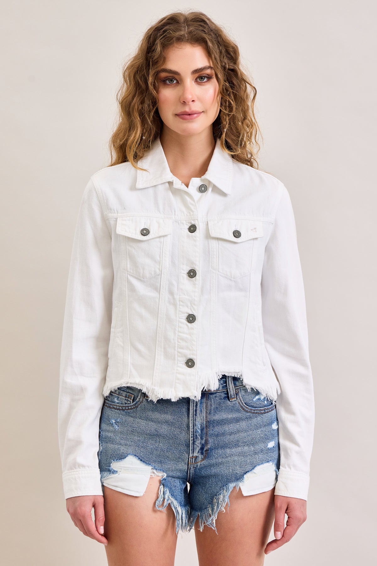 A woman with wavy hair wears the Raw Hem Denim Jacket by Hidden Jeans over distressed blue denim shorts, standing against a neutral background.