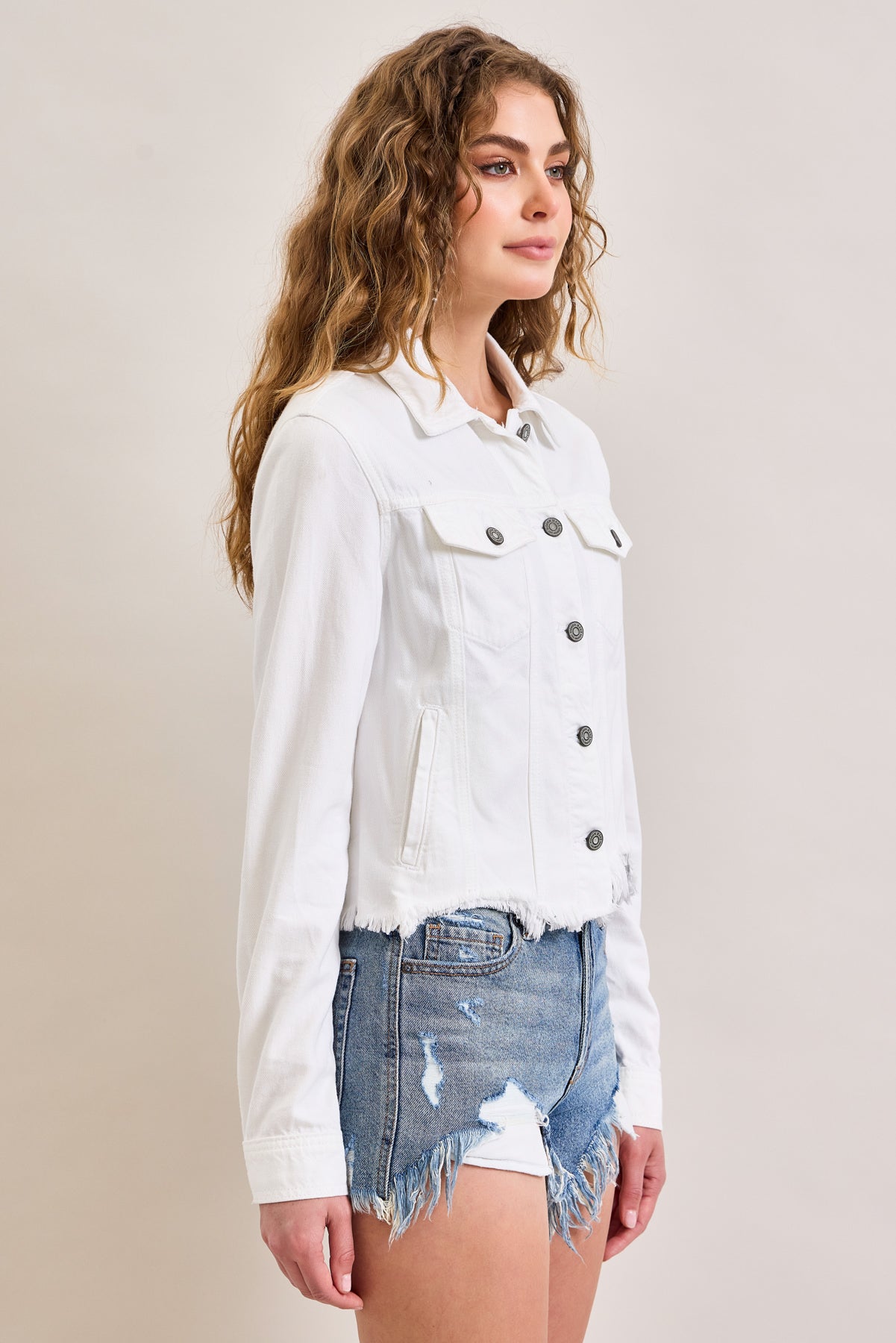 A woman with wavy hair wears the Hidden Jeans Raw Hem Denim Jacket, fitted in white with black buttons, paired with distressed blue denim shorts. She stands against a plain light background, facing slightly to the right.
