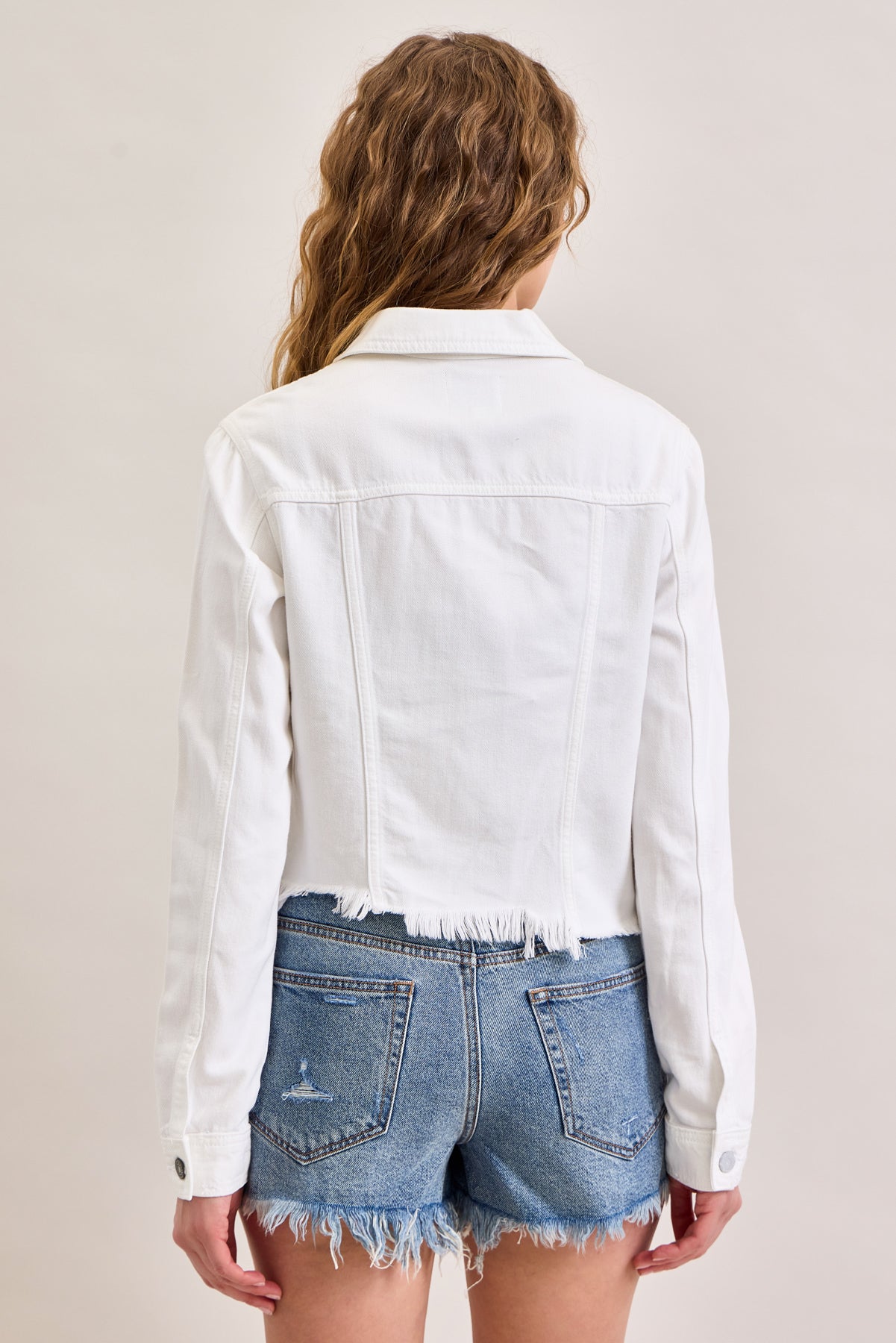 A person with wavy, light brown hair faces away from the camera, wearing Hidden Jeans' Raw Hem Denim Jacket in white and light blue distressed shorts, against a plain, light background.