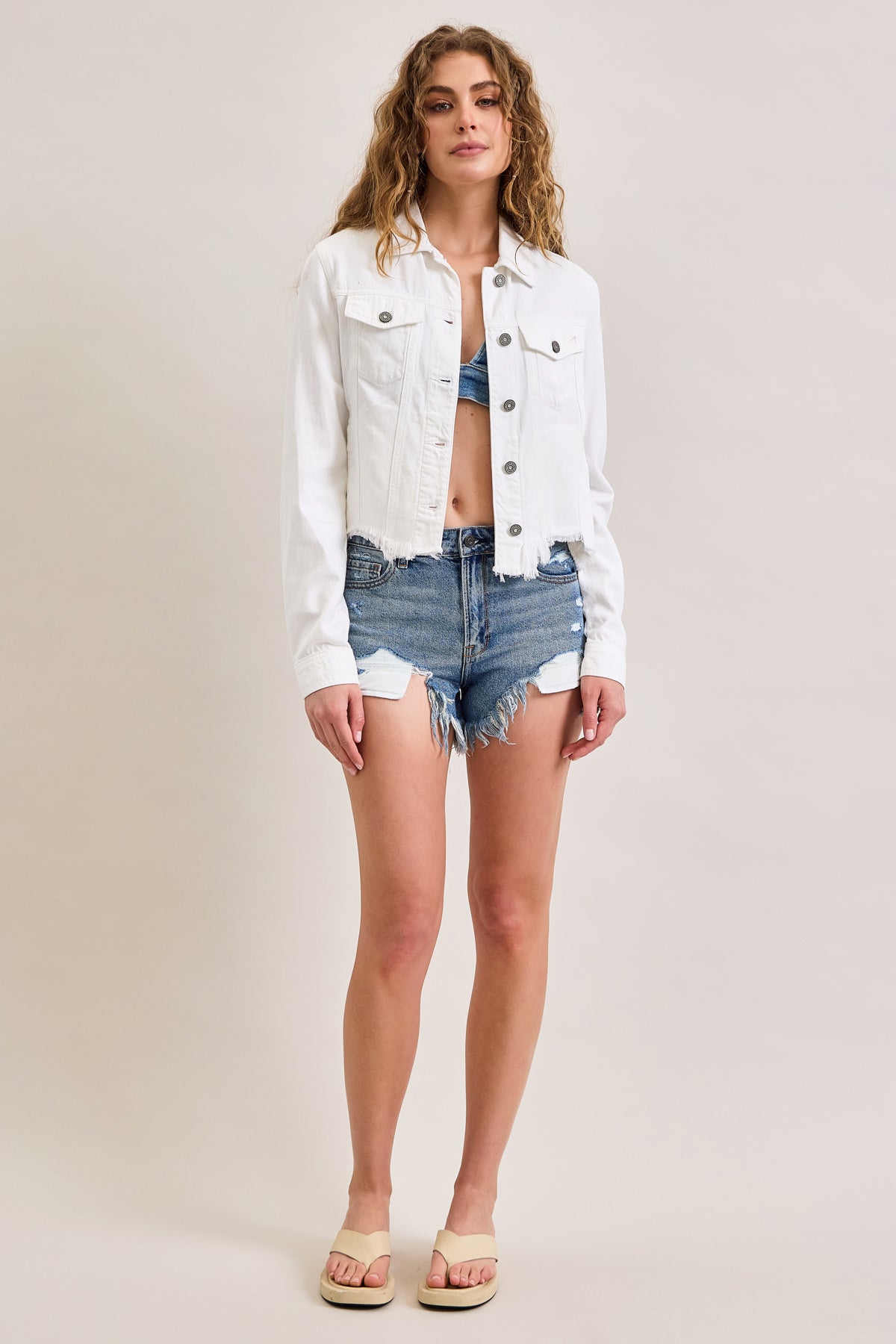 A woman with long, wavy hair and a relaxed expression wears the Hidden Jeans Raw Hem Denim Jacket over a blue bralette, paired with distressed denim shorts and beige sandals, standing against a plain background.