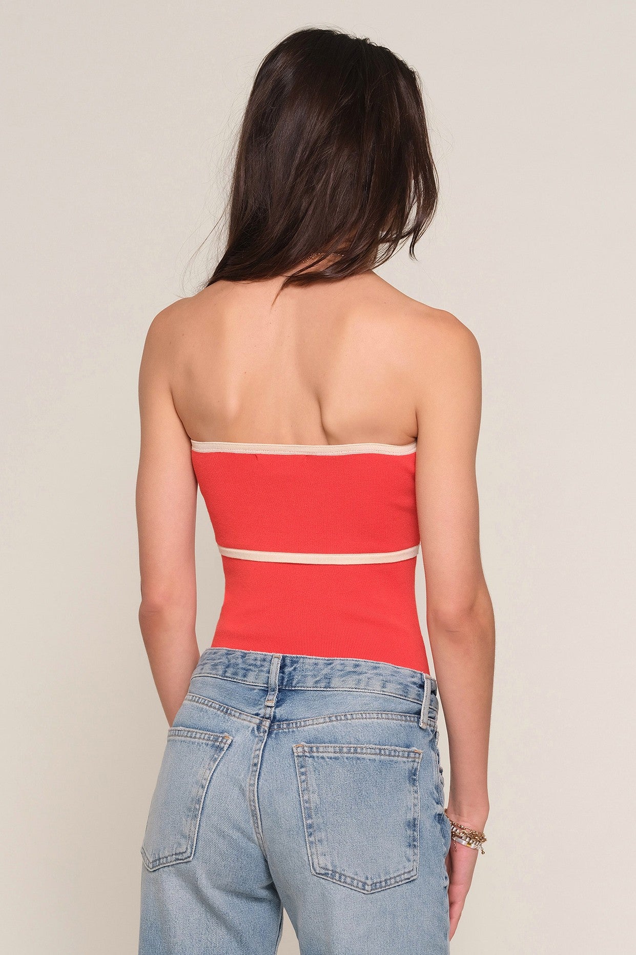 A woman with long brown hair, seen from behind, wears the Heartloom Avril Top—a slim fit strapless red top with beige contrast piping—paired with light blue high-waisted jeans and a bracelet on her left wrist.