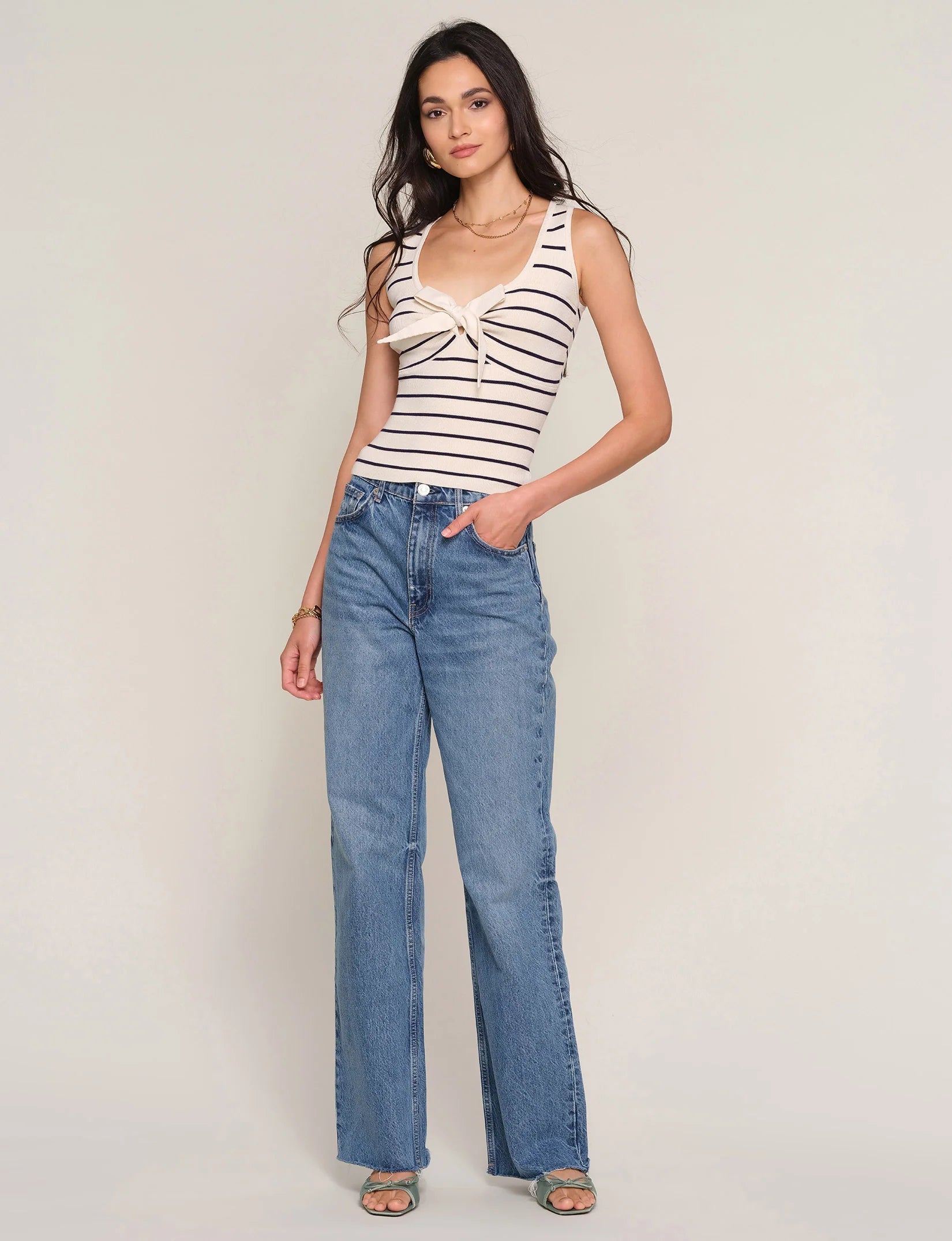 A woman with long wavy hair poses confidently against a light background, wearing Heartloom's Tiffy Top—a slim fit, striped sleeveless top with a front tie—paired with high-waisted blue jeans and green sandals.