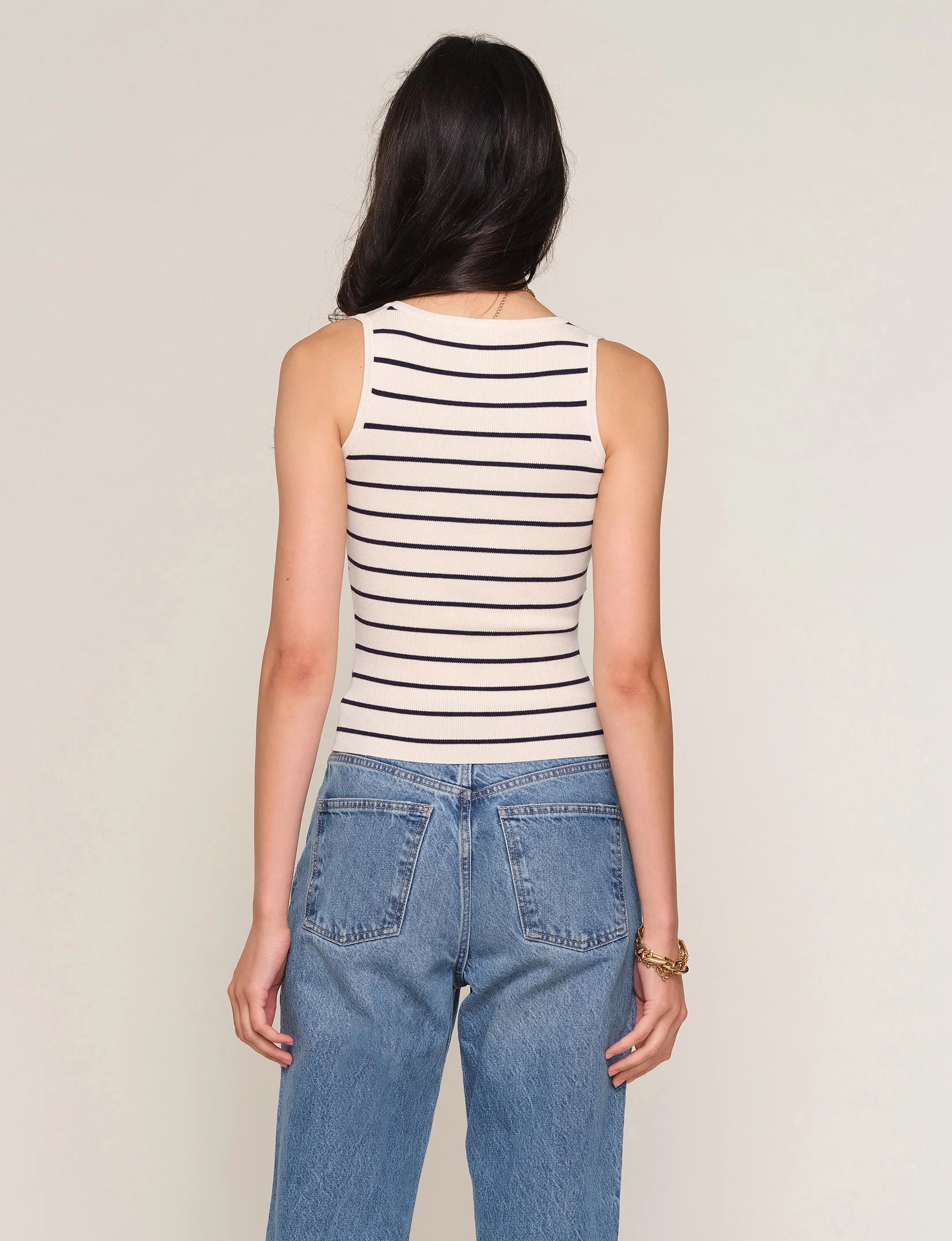A person with long dark hair is seen from behind, wearing the Heartloom Tiffy Top—a slim-fit, sleeveless white tank with black stripes—paired with blue jeans against a plain light background.