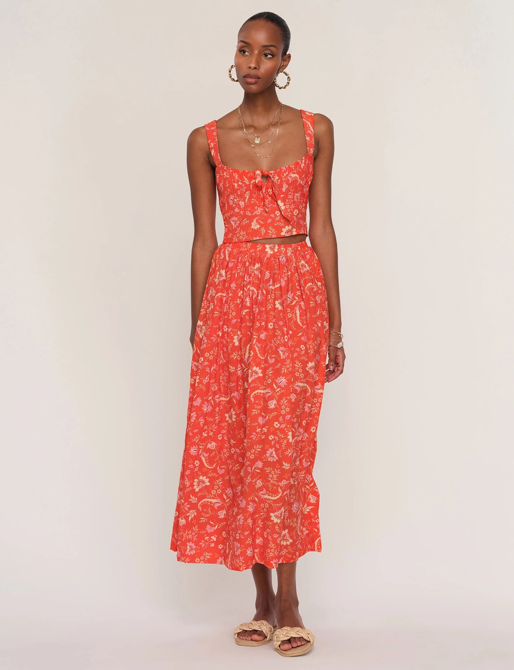 A woman wears the Heartloom Camille Set Tank— a sleeveless red crop top with a matching floral high-waisted flared skirt—paired with hoop earrings, layered necklaces, and sandals, standing against a plain light background.
