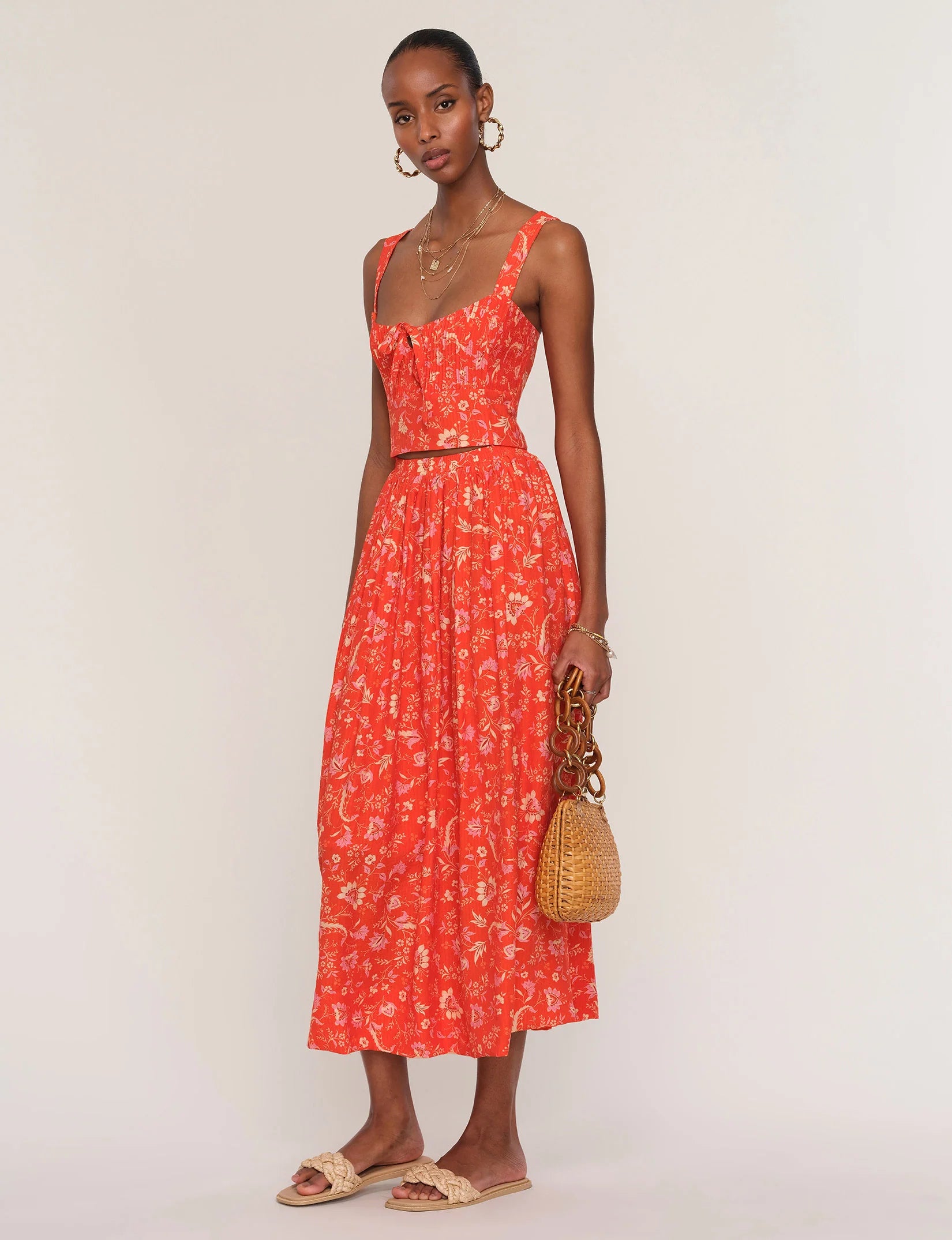 A woman wearing Heartloom’s red Camille Set Skirt stands against a plain light background, styled with beige slide sandals, hoop earrings, layered necklaces, and holding a woven handbag.