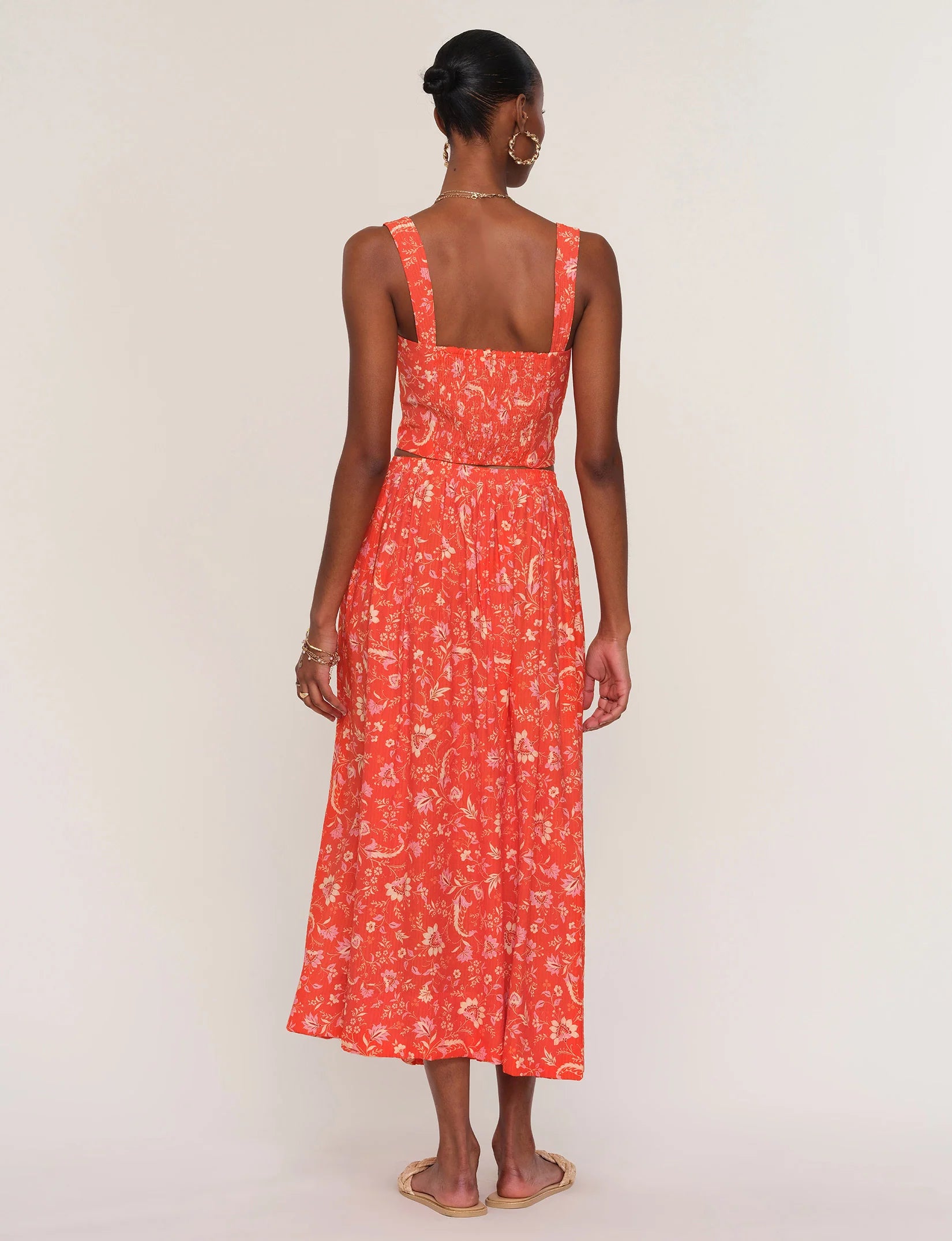 A woman stands with her back to the camera in the Heartloom Camille Set Skirt, an ankle-length red floral piece with a smocked back, styled with flat sandals and a neat low bun against a plain light background.