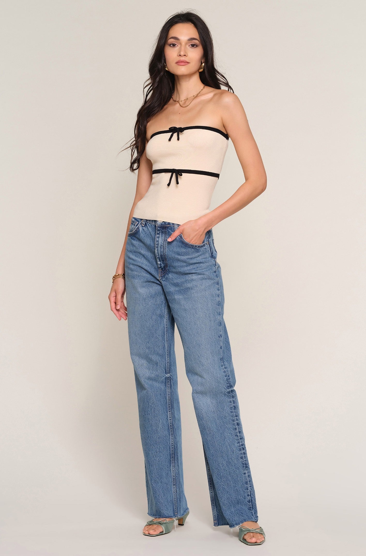A woman with long wavy hair stands against a plain background wearing the Heartloom Avril Top in beige rib knit with black trim and bows, paired with blue slim fit high-waisted jeans and green heeled sandals, one hand in her pocket.