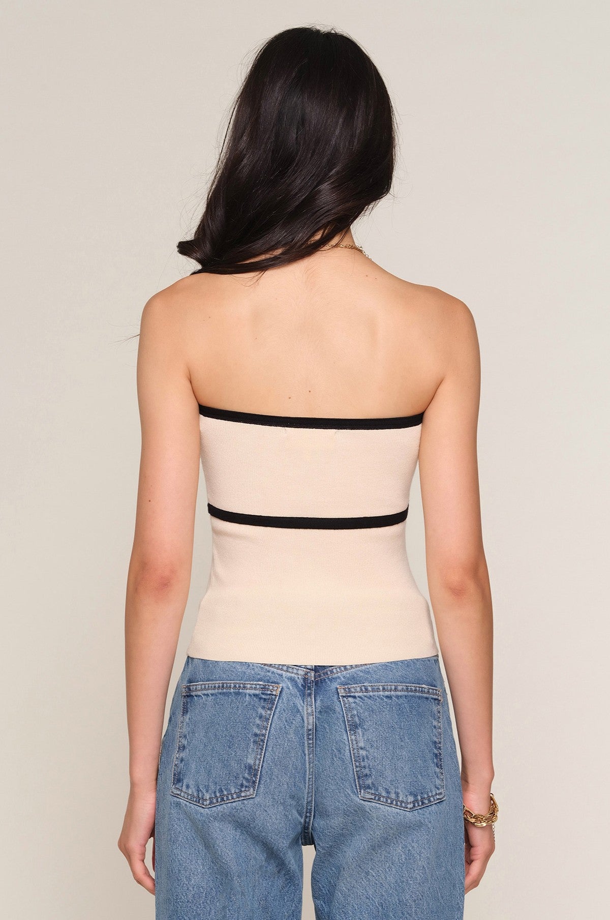 A woman with long dark hair, seen from the back, wears the strapless Avril Top by Heartloom in a rib knit slim fit, paired with high-waisted blue jeans. She stands against a plain light background.