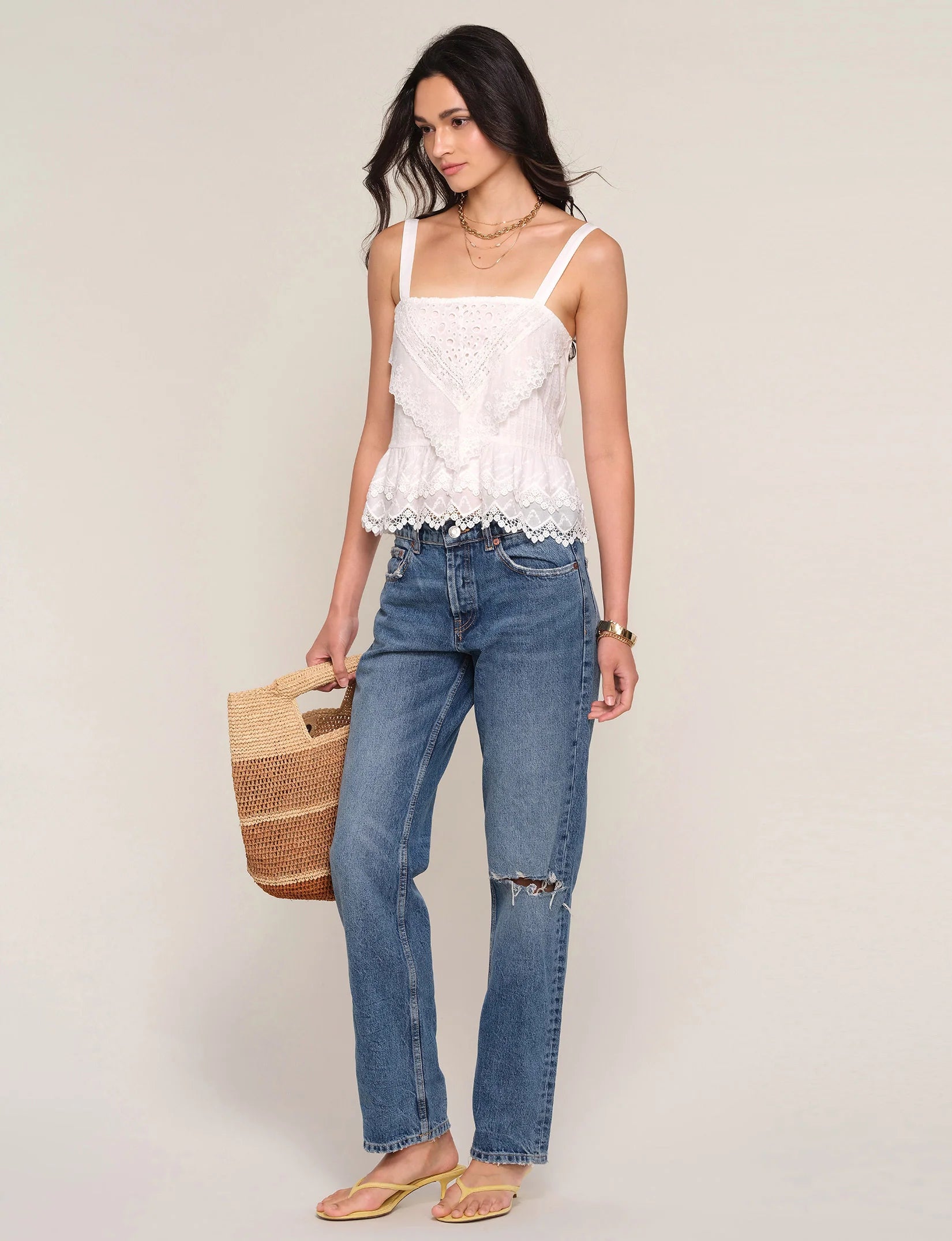 A woman wears the Heartloom Fernando Top, a white lace detail sleeveless style, paired with blue ripped jeans and yellow sandals. She holds a woven straw bag, has long dark hair and layered necklaces, standing against a plain light background.