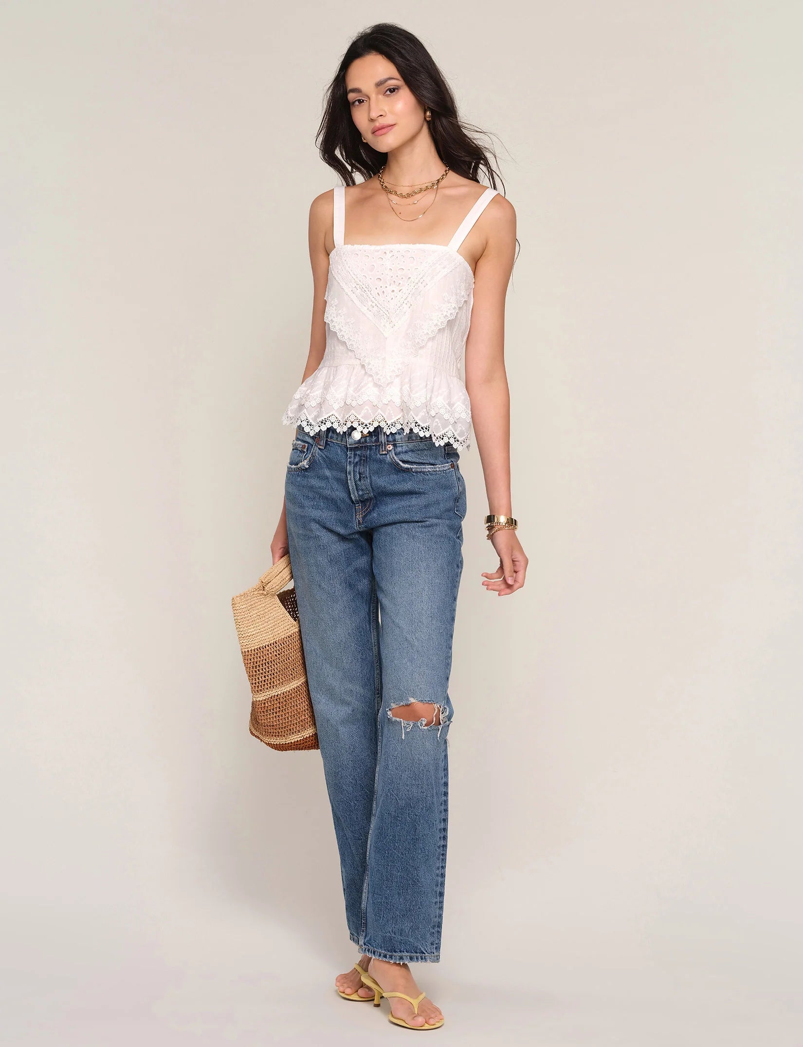 A woman wears the Heartloom Fernando Top in ivory—a lace detail camisole with adjustable straps—paired with blue ripped jeans, sandals, a woven bag, layered necklaces, and a bracelet, standing against a plain background.