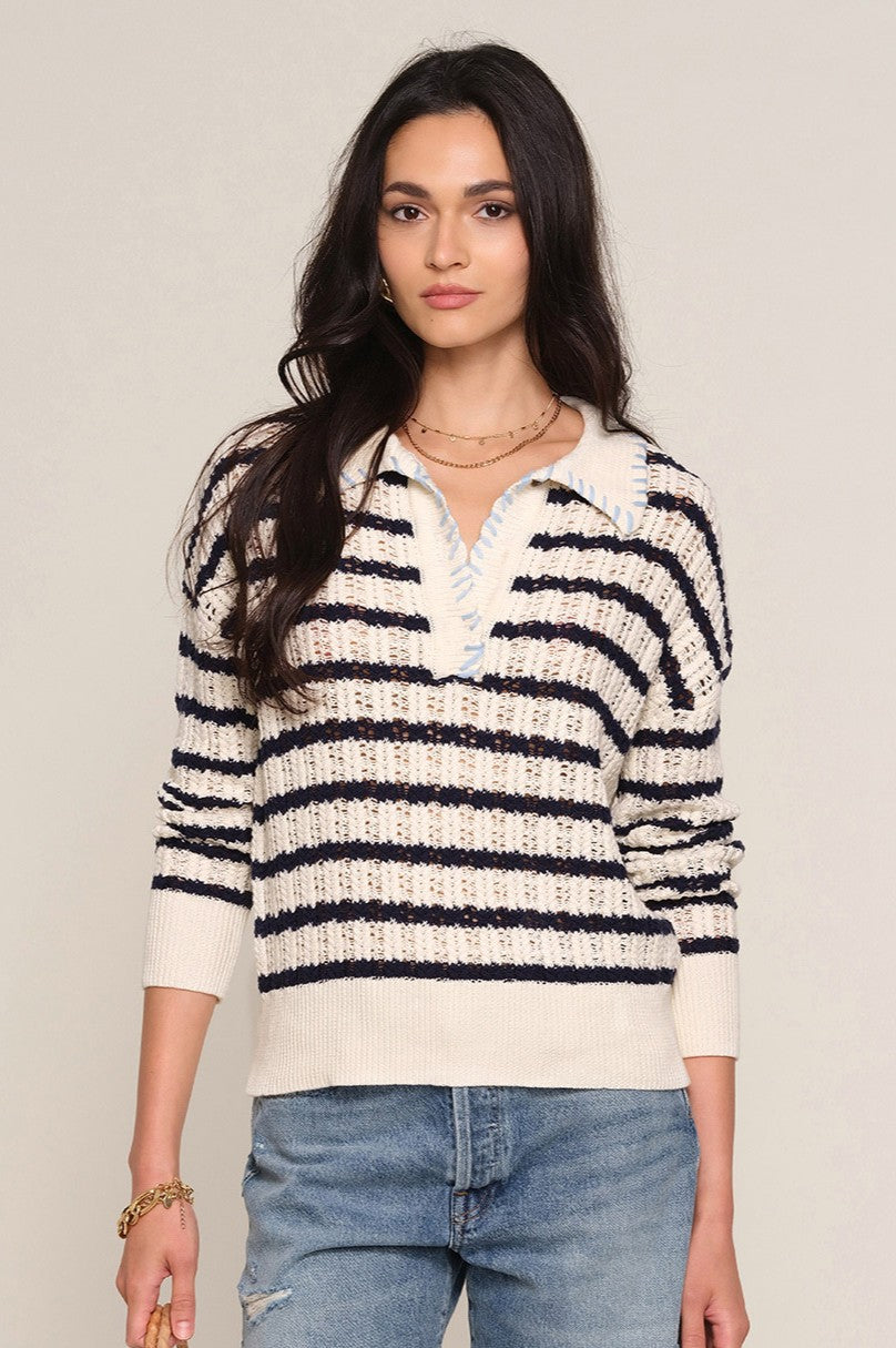 A woman wears the Heartloom Isme Sweater, a relaxed 100% cotton polo in cream and navy stripes with a collared neckline and light blue stitching, pairing it with ripped denim jeans and gold jewelry.
