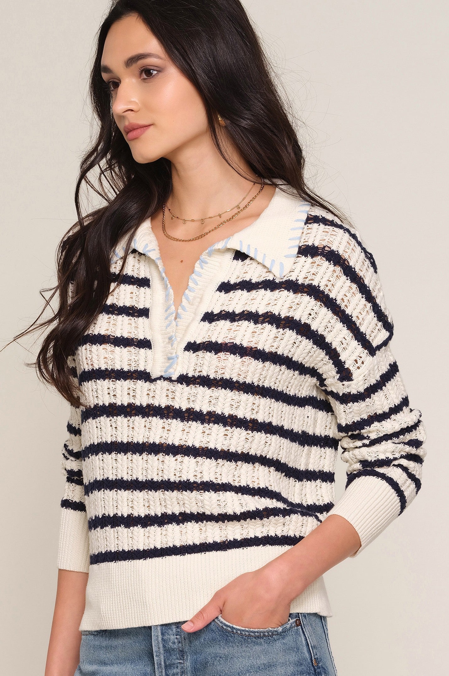 A woman with long dark hair wears the Heartloom Isme Sweater, a relaxed 100% cotton polo featuring cream and navy stripes, a collared neckline, and light blue accents. She stands sideways in blue jeans, looking to the left.