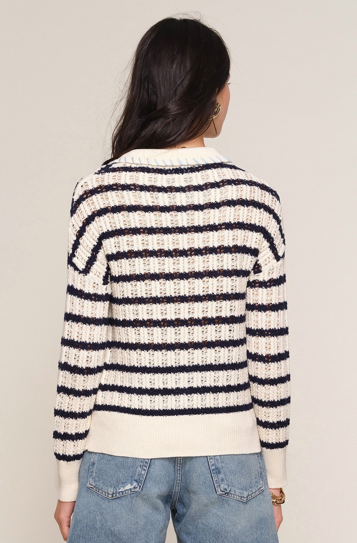 A person with long dark hair is seen from behind wearing the Heartloom Isme Sweater—a cream and navy striped 100% cotton knit with a relaxed polo fit—paired with light blue jeans against a plain background.