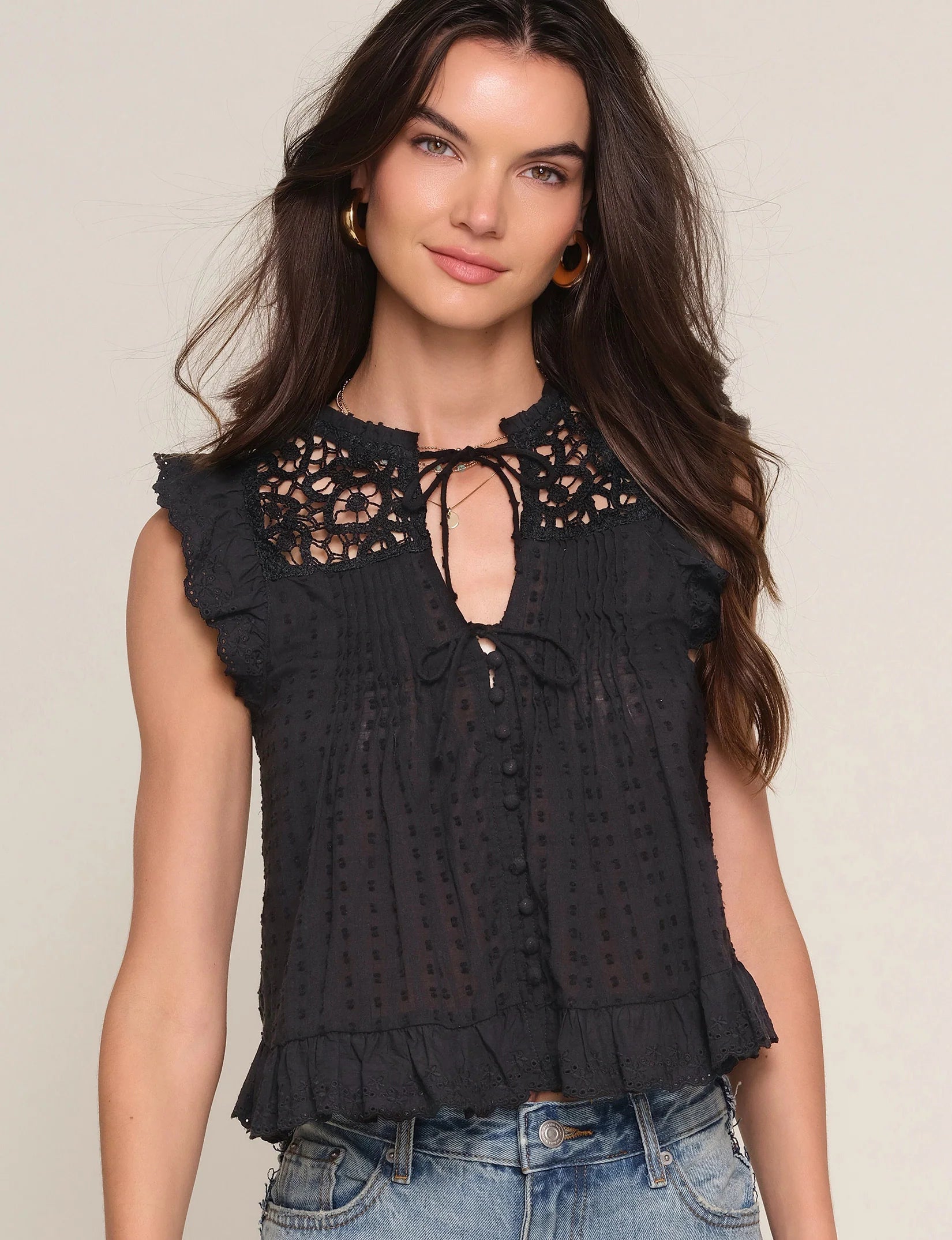 A woman with long brown hair wears the Heartloom Rayna Top, a sleeveless black blouse with lace yoke, tie-front, and ruffled edges, paired with relaxed fit blue denim jeans, standing against a plain light background.