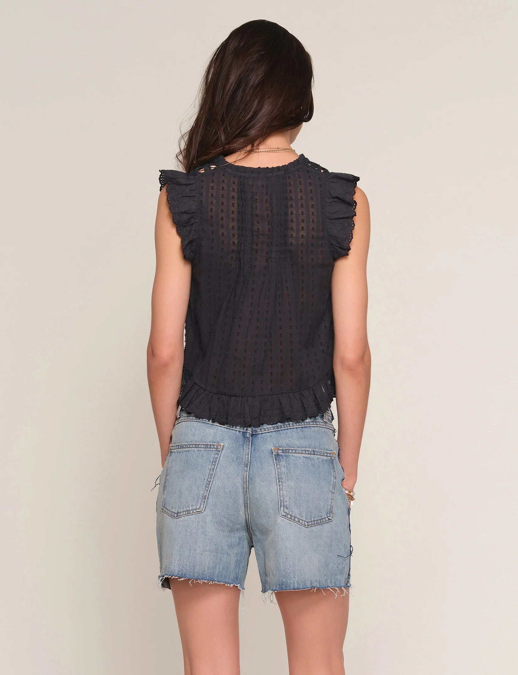 A woman with long brown hair stands with her back to the camera, wearing the Heartloom Rayna Top—a relaxed fit, sleeveless black eyelet top with ruffled edges—and light blue denim shorts with frayed hems against a plain background.