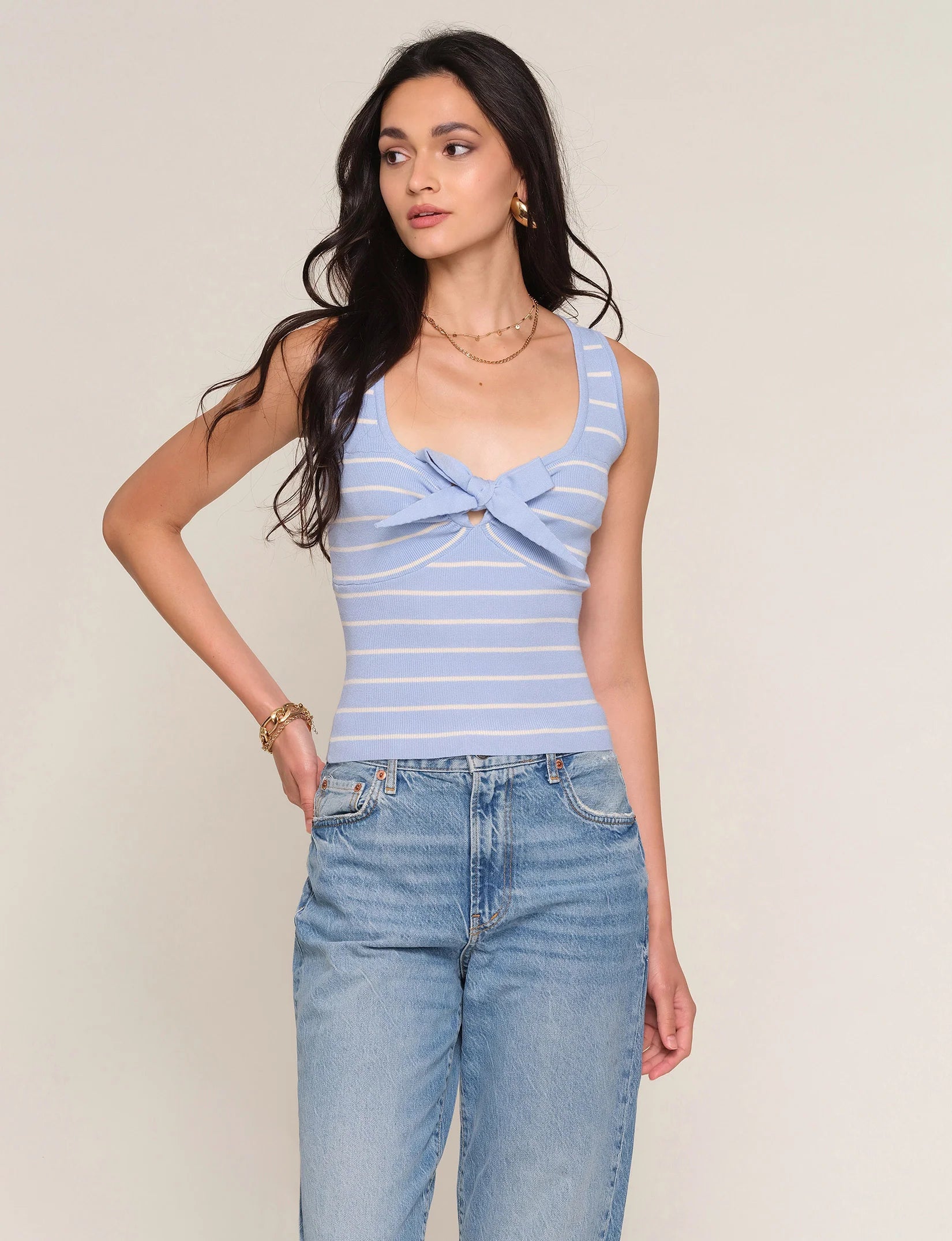A woman with long dark hair wears the Heartloom Tiffy Top—a slim-fit, light blue and white striped style with a front knot—paired with high-waisted jeans and gold jewelry, standing against a plain light background.